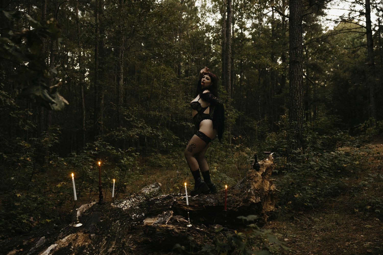 Woman in black bodysuit posing among candles in a dark moody forest.