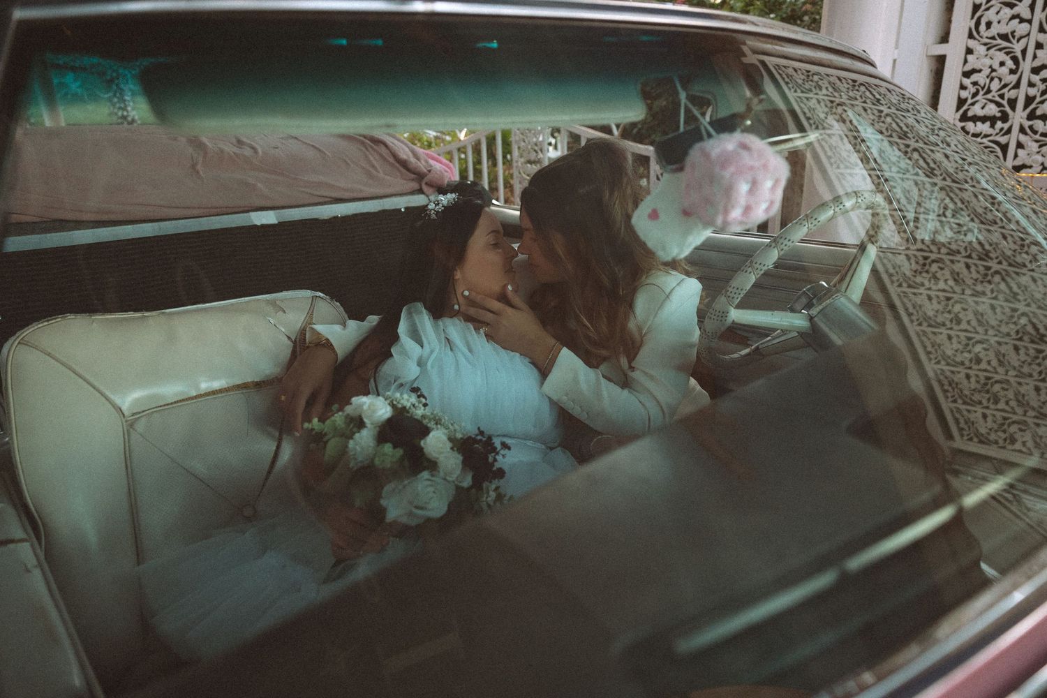 Bride and groom share a kiss inside a vintage classic car with white leather interior.