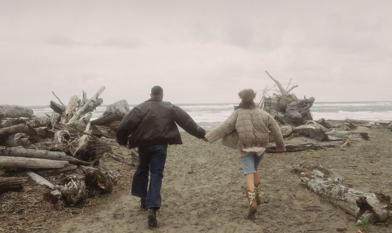 Couple holding hands walking along a driftwood-lined beach on a cloudy day.