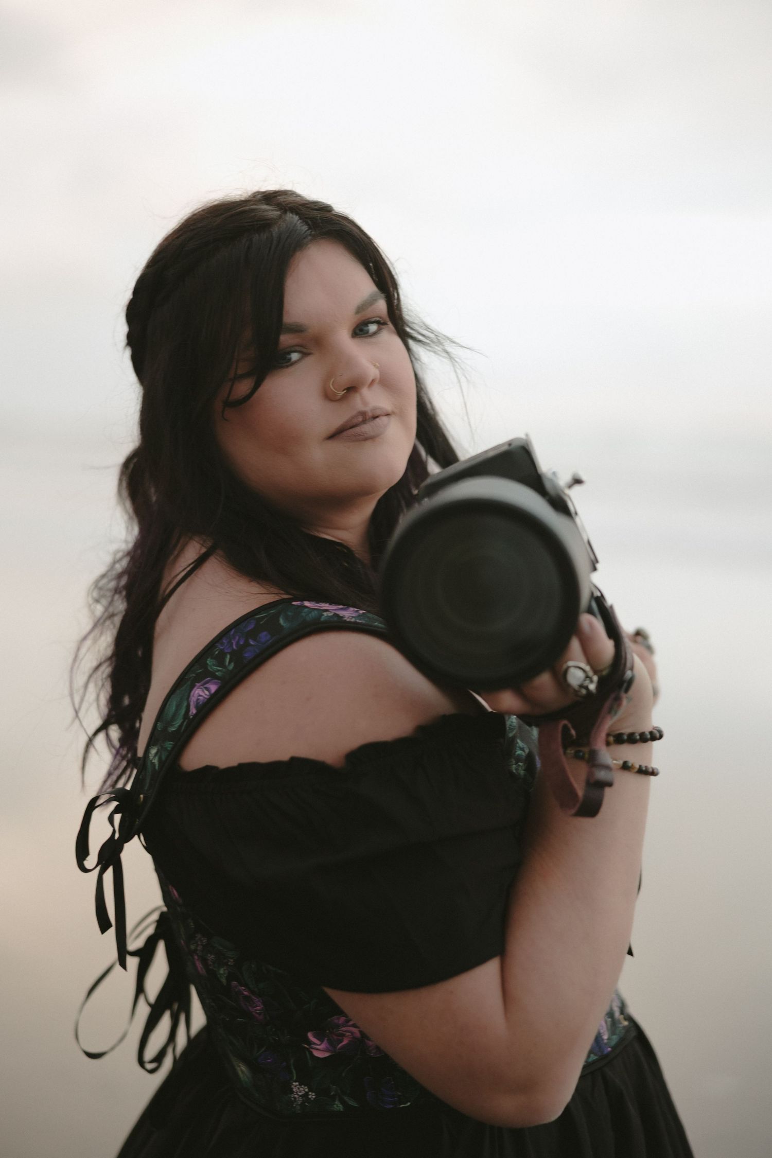 A brunette woman in a black off-shoulder dress holds a camera at golden hour near water.