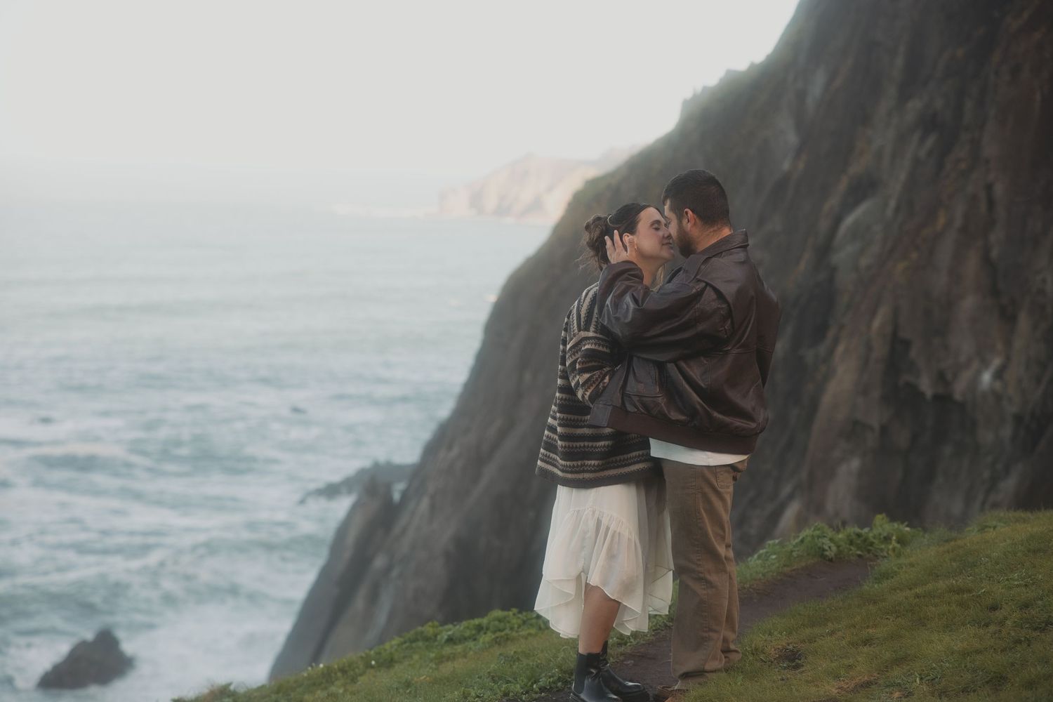 Couple shares a tender kiss on a misty coastal cliff edge with crashing ocean waves below.