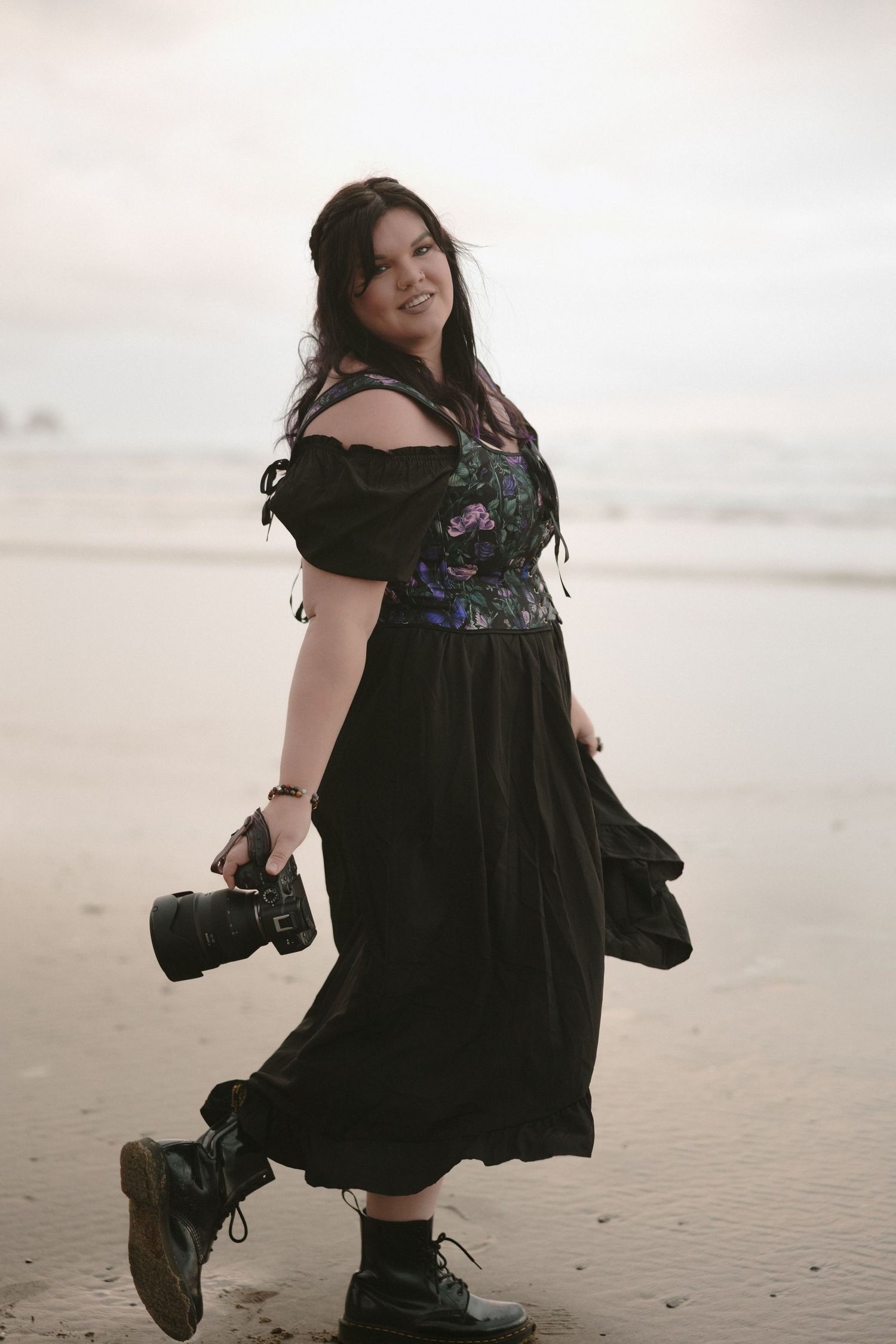 Woman in black off-shoulder dress walking on beach holding a camera.