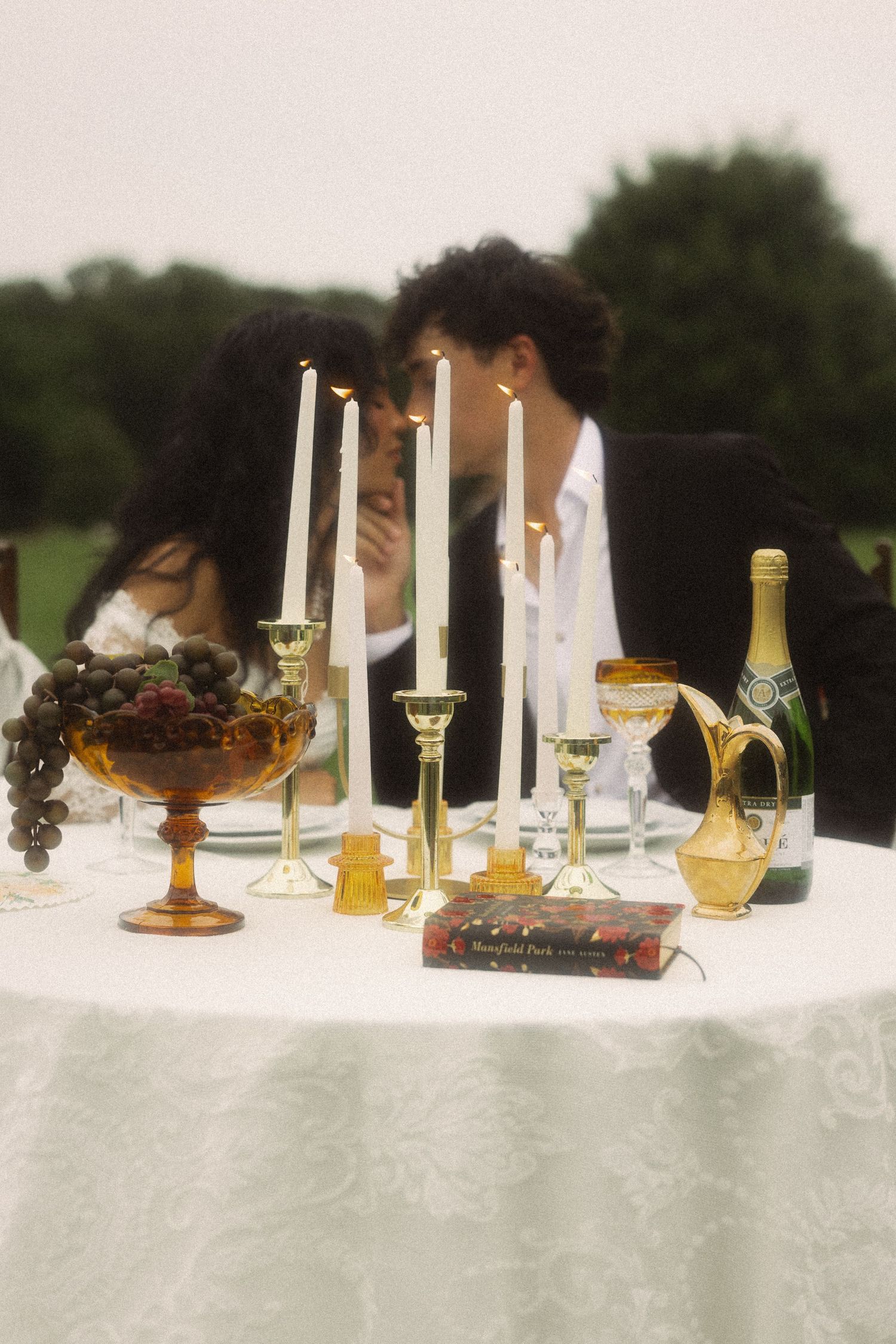 Couple leans in for a kiss behind an elegant outdoor candlelit table set with champagne and fruit.