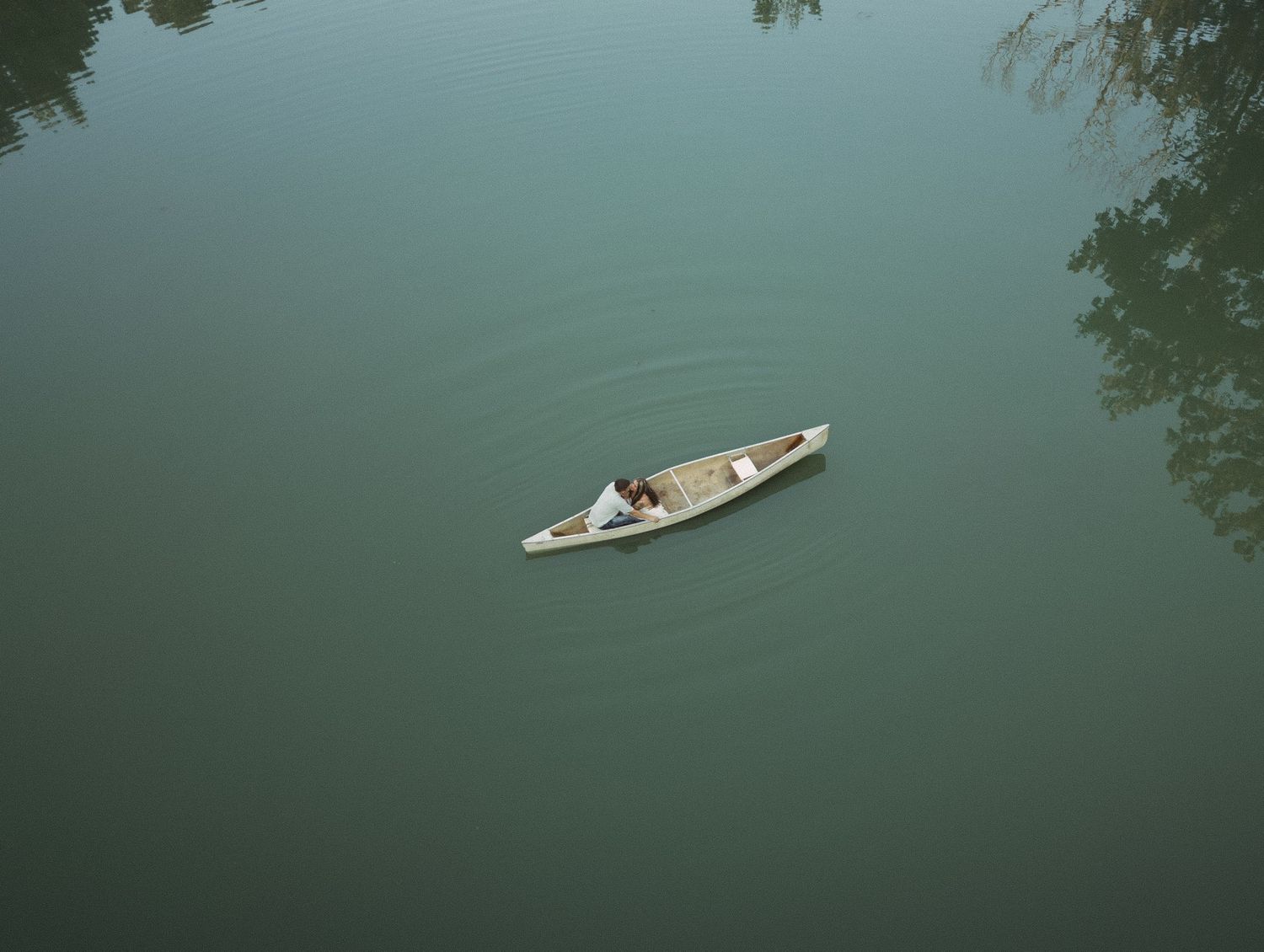 Aerial view of a person paddling a small white canoe across calm green water near tree-lined shore.