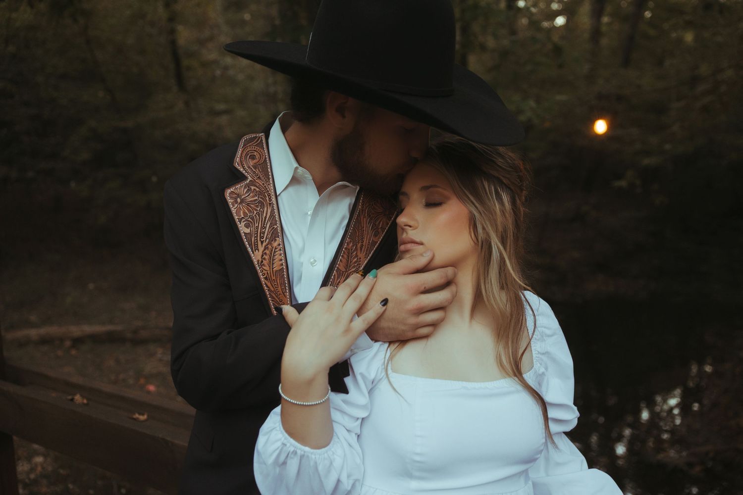 Couple in western attire sharing an intimate embrace near a forest bridge at dusk.