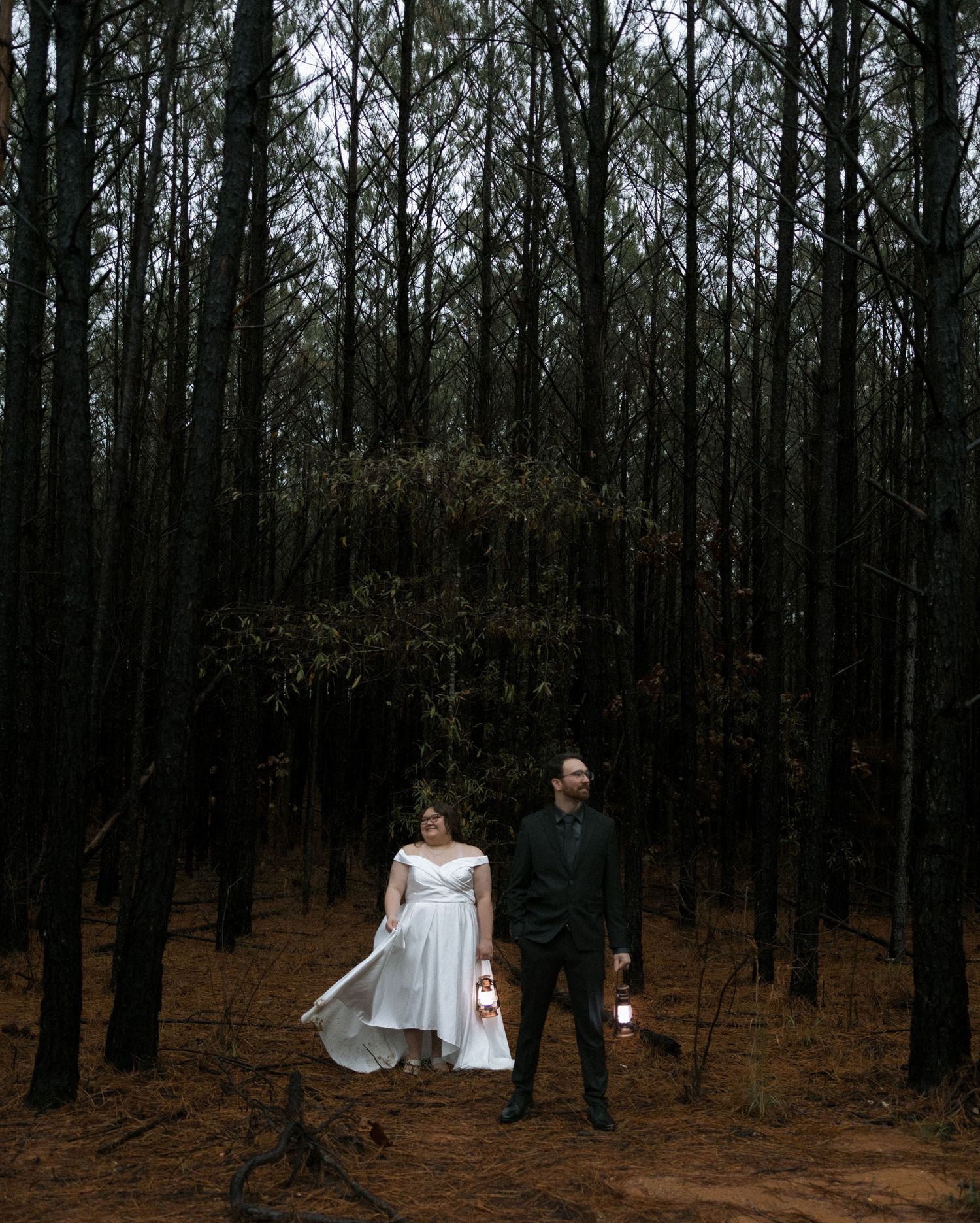 A bride and groom stand apart in a dark pine forest, the bride holding her white gown train.