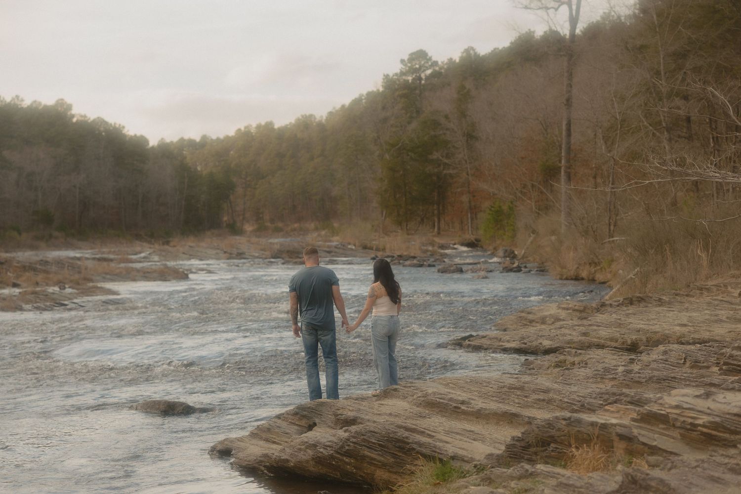 Couple holding hands while standing on rocks beside a scenic river in a forest.