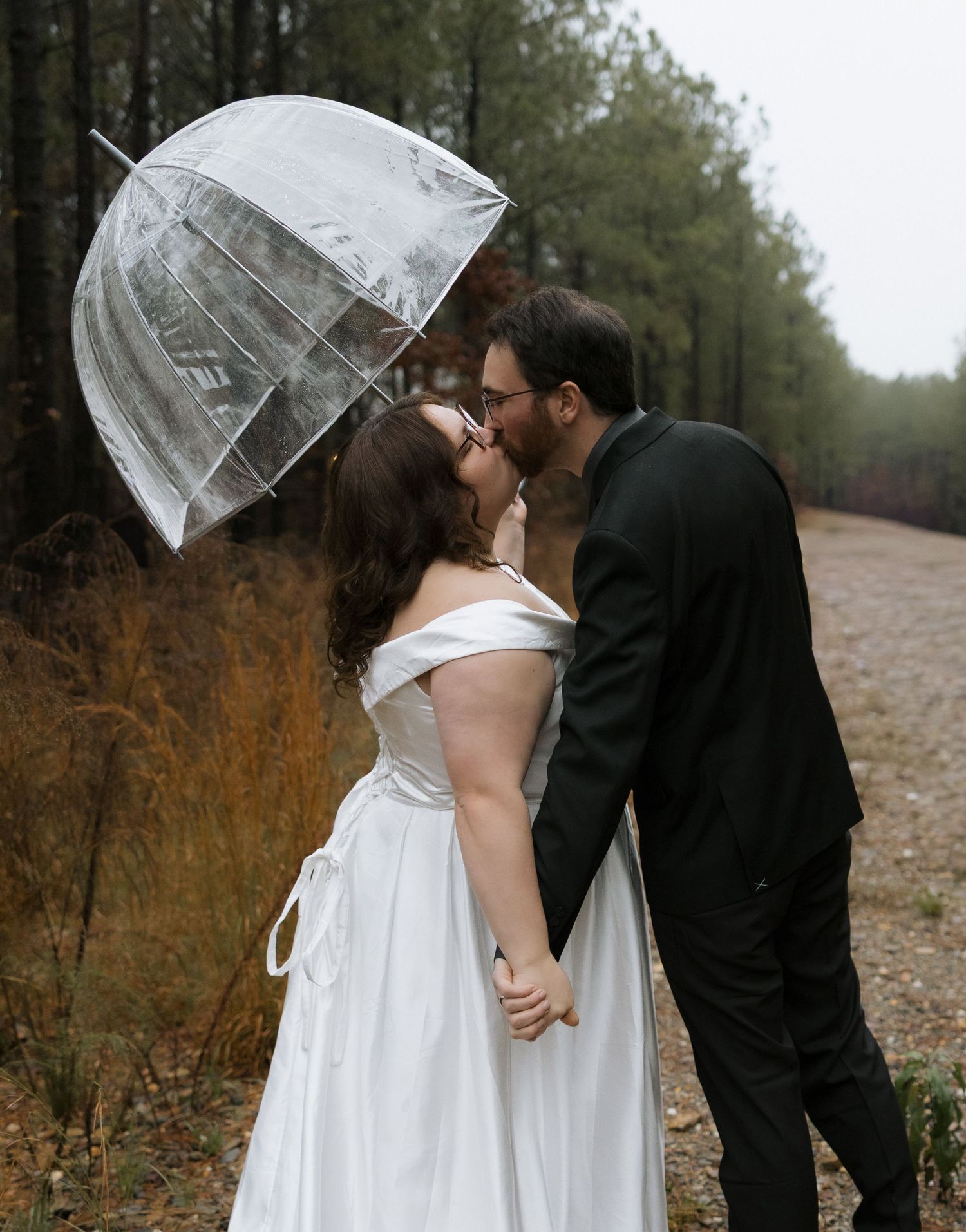 Bride and groom sharing a romantic kiss under a clear bubble umbrella on a rainy wedding day outdoors.