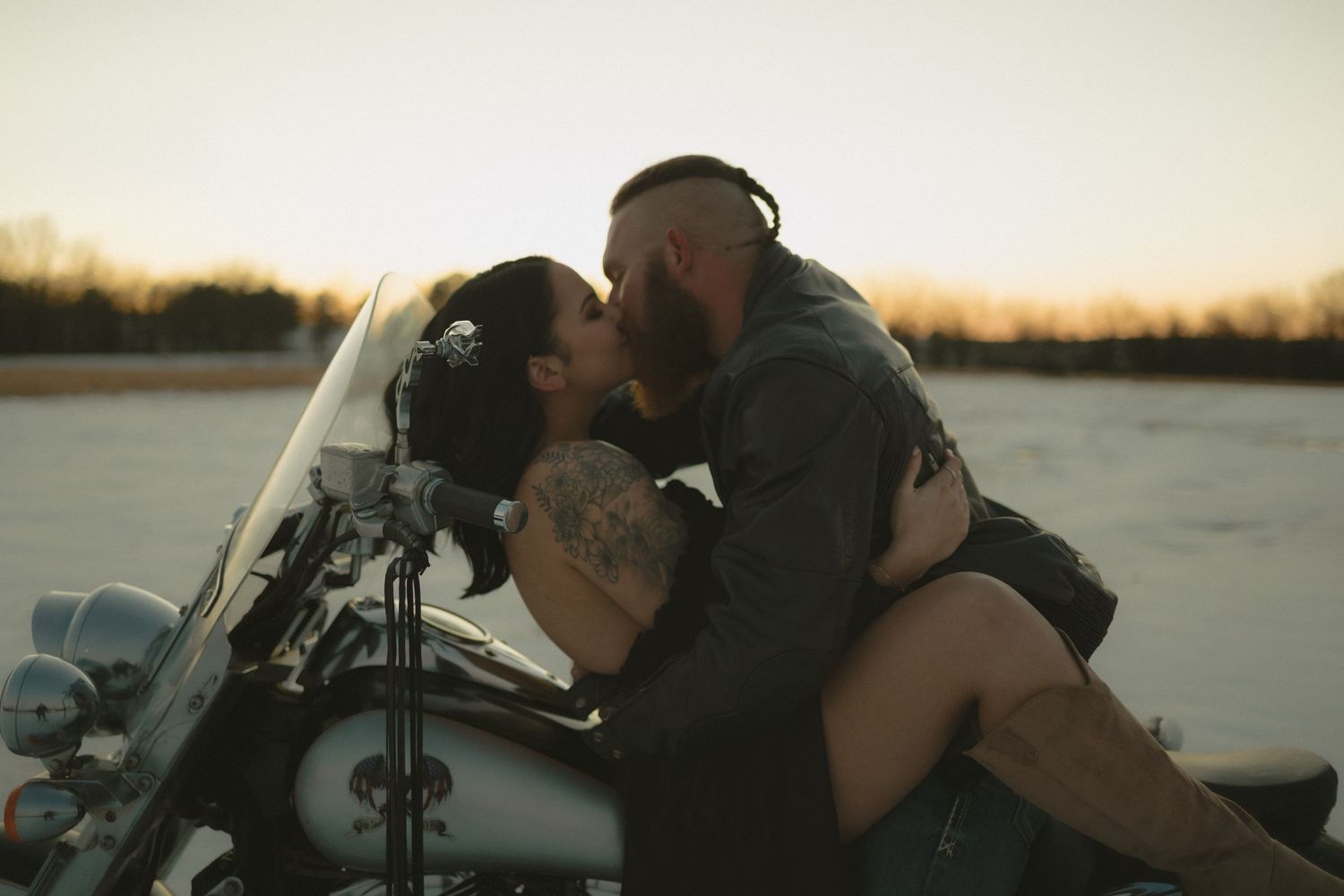 Tattooed couple sharing a passionate kiss on a motorcycle at sunset.