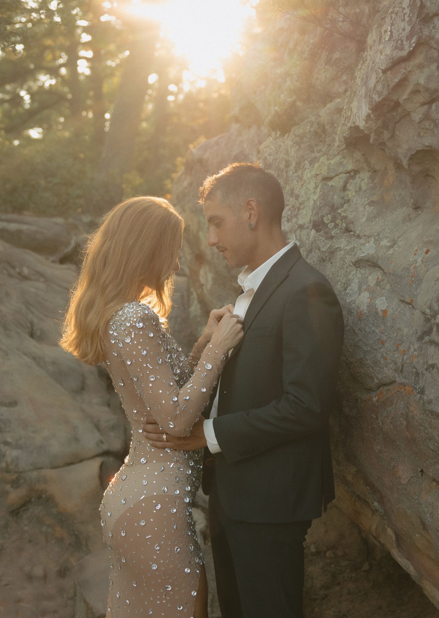 Couple embracing in golden sunlight against rocky cliff, woman in embellished gown.