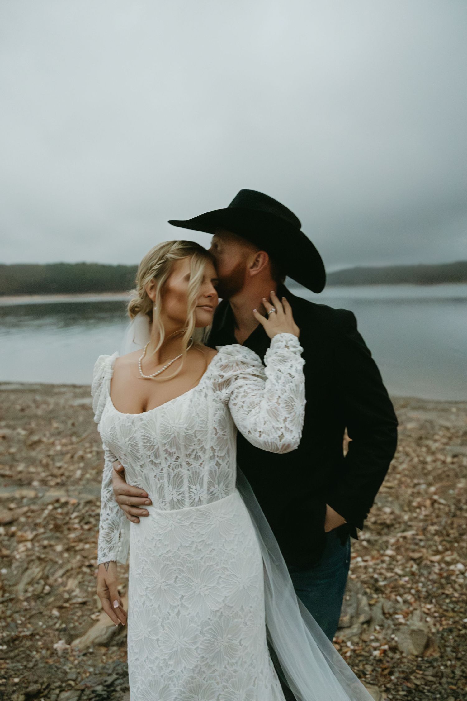 Bride in lace gown and groom in cowboy hat sharing a kiss by a lake.