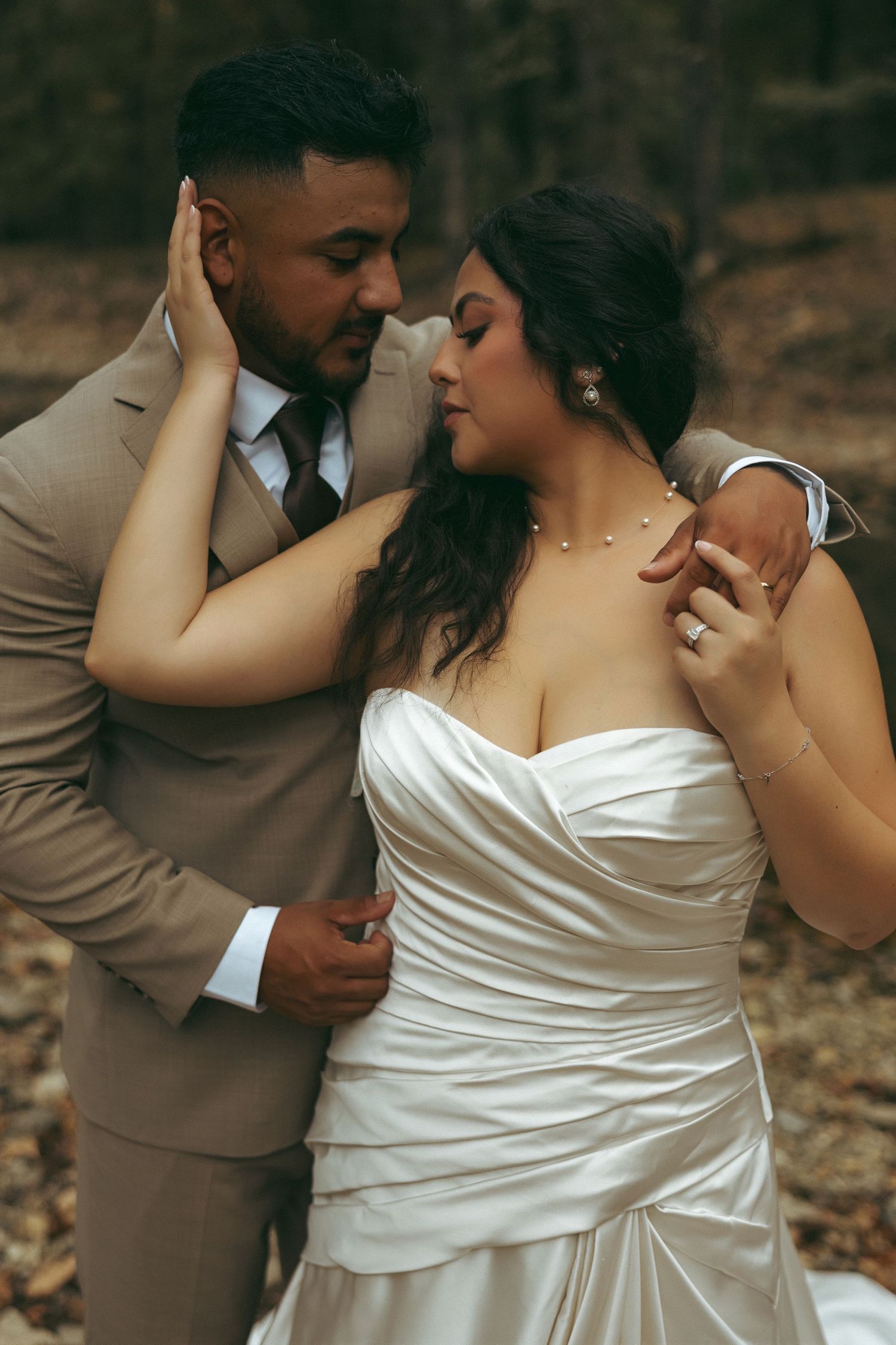 A bride in a strapless satin gown and groom in a tan suit share a tender moment in an autumn forest.