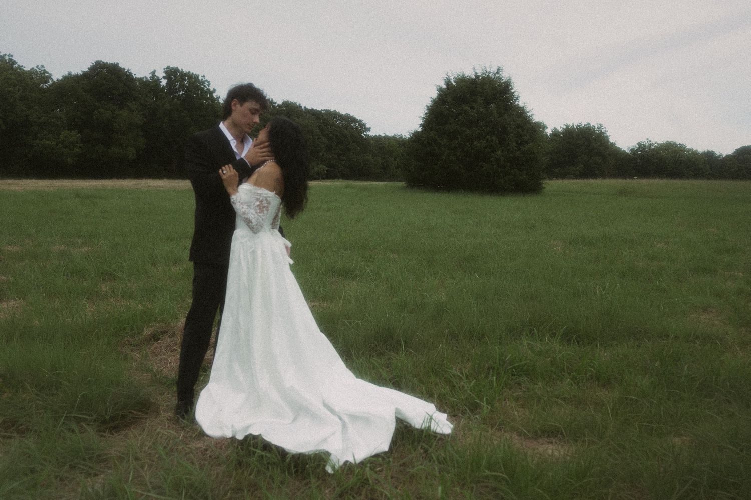 A bride and groom embrace in a lush green meadow, the bride's white gown trailing on the grass.