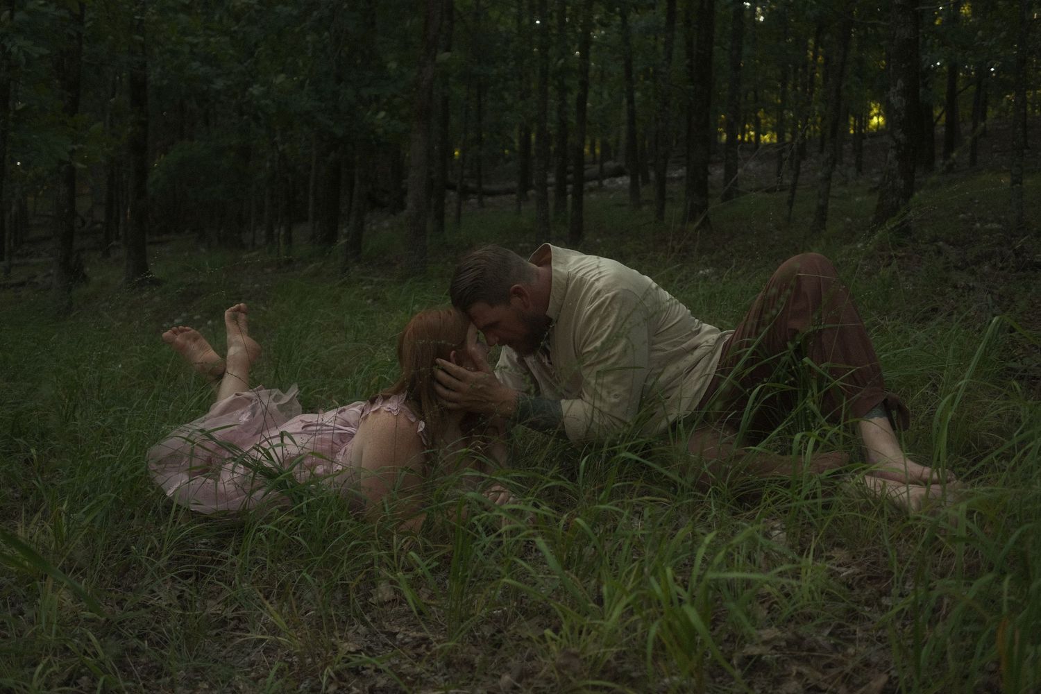 Couple sharing a romantic kiss while lying in a lush green forest clearing at dusk.