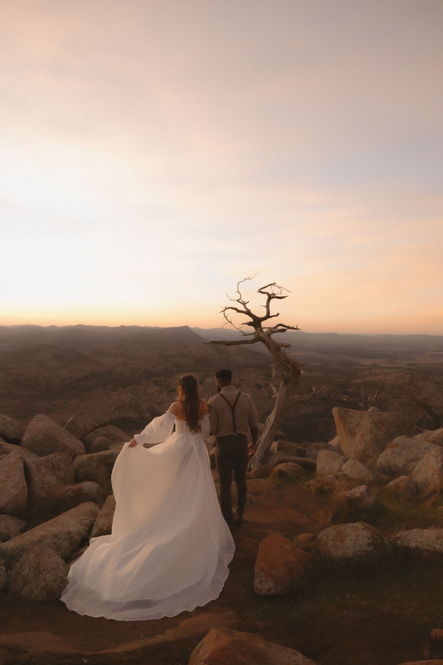 Bride and groom standing on rocky mountaintop at sunset with flowing white dress.