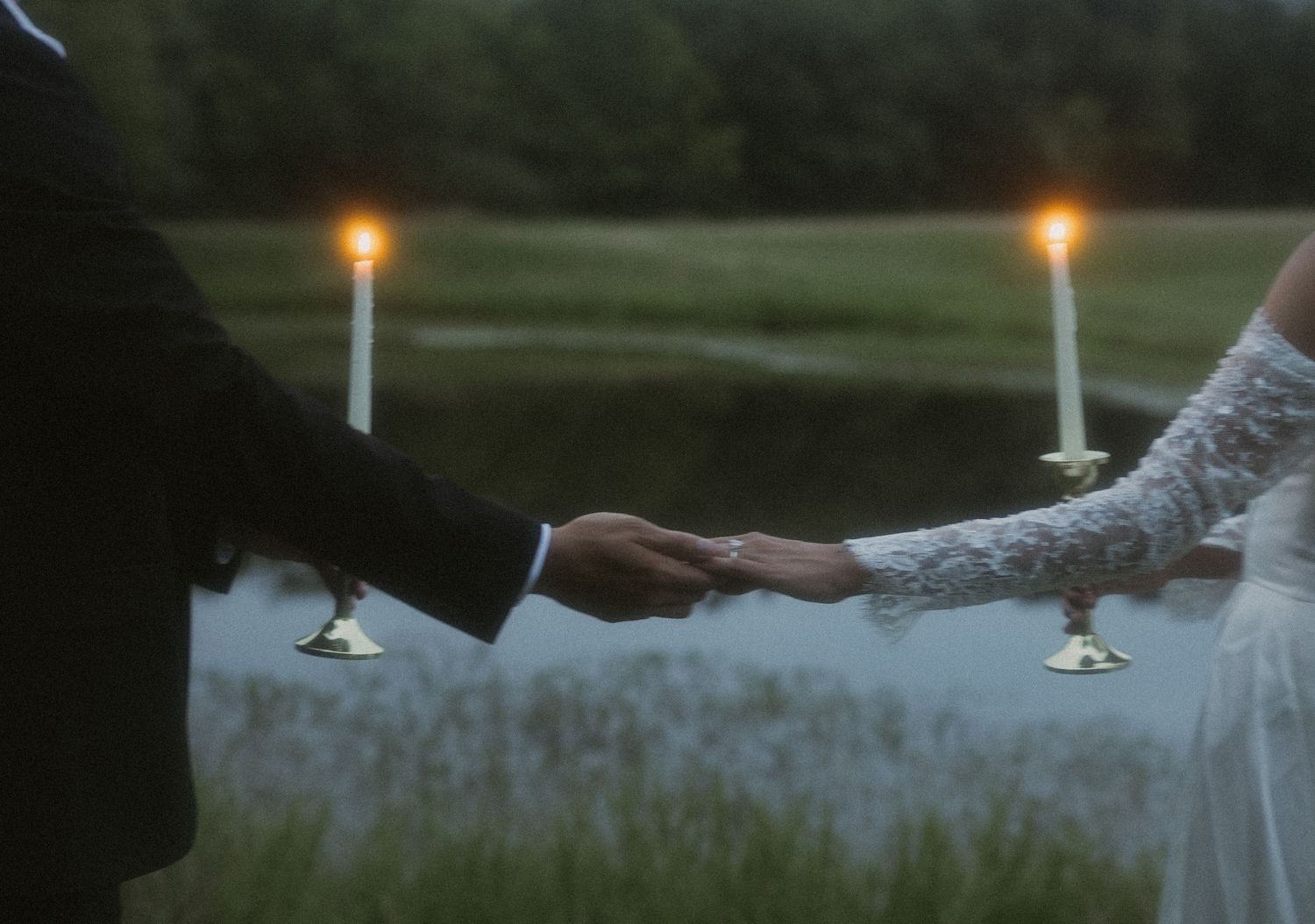 Bride and groom holding hands by candlelight near a lake at dusk.