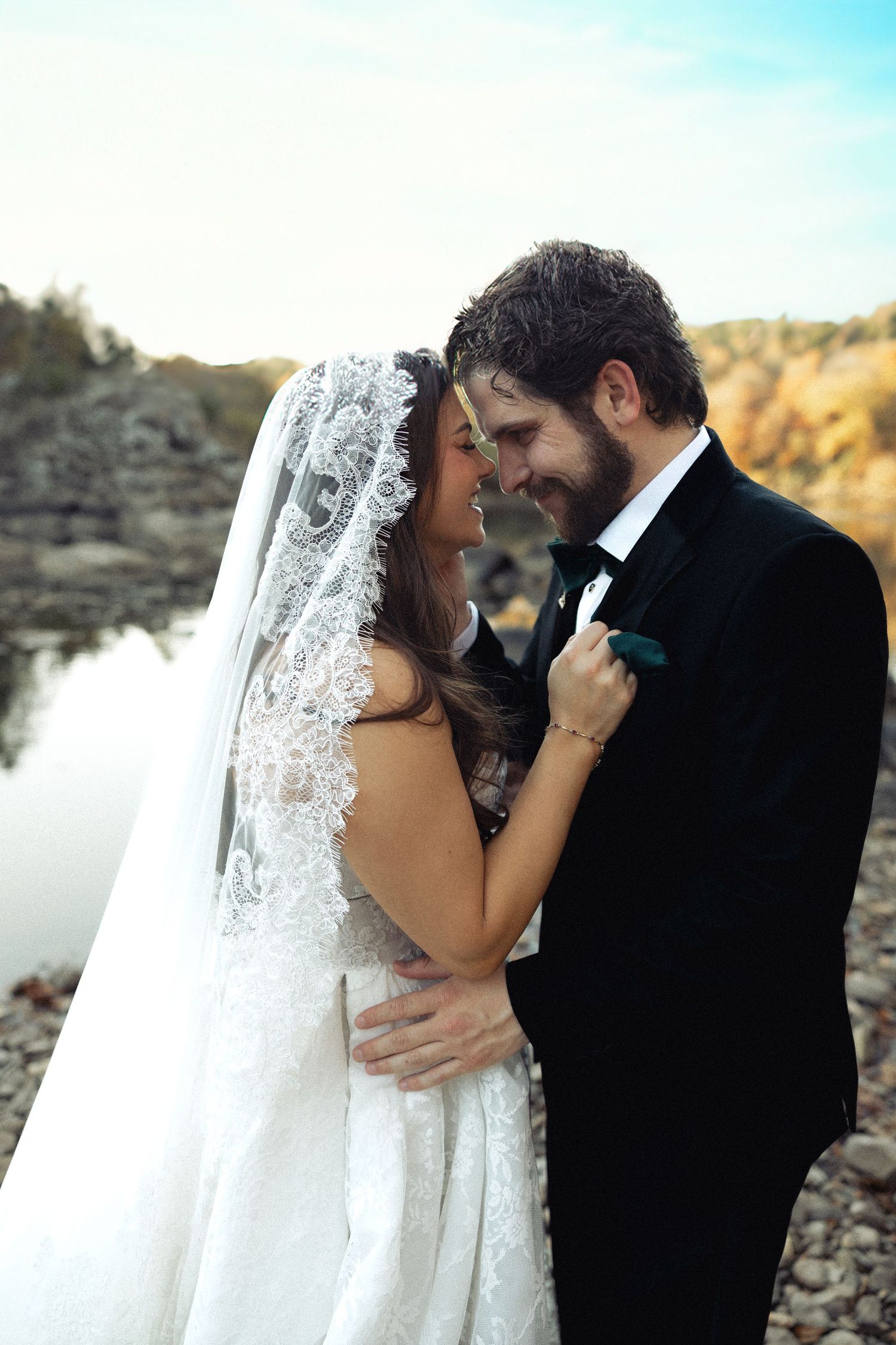 Smiling bride in lace veil and groom in tuxedo embracing near a scenic rocky waterway.