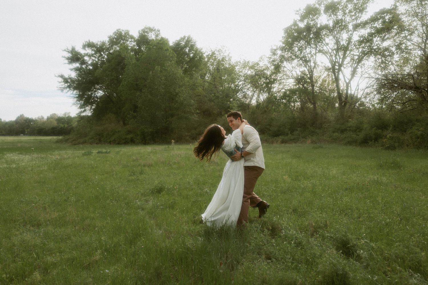 Couple sharing a romantic dance and kiss in a green open field with trees behind.