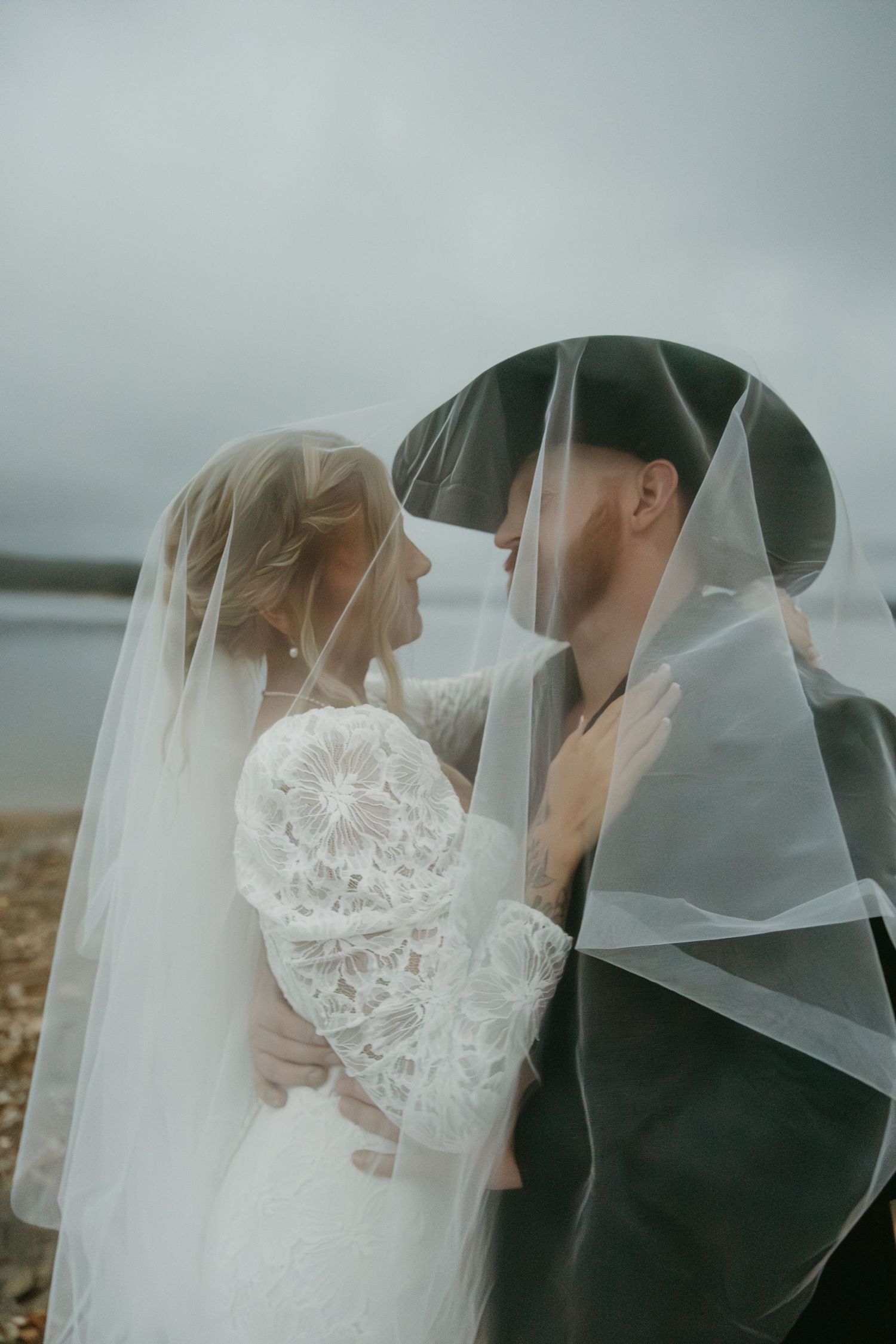 A bride and groom share an intimate moment under a flowing veil by a lakeside on a cloudy day.