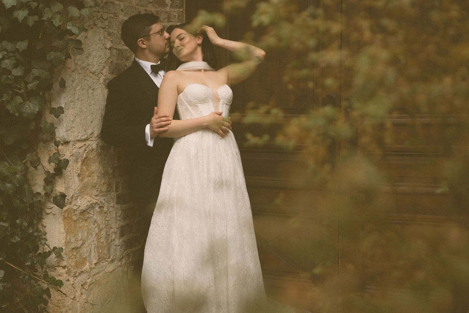 Couple in wedding attire sharing an intimate moment against a rustic stone wall surrounded by greenery.