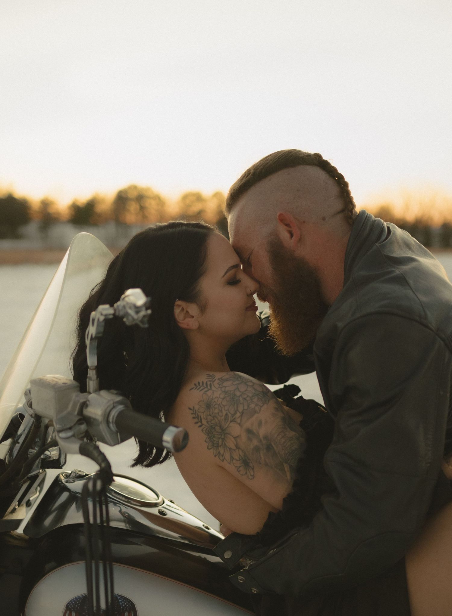 Tattooed couple sharing romantic kiss on vintage motorcycle at golden sunset.
