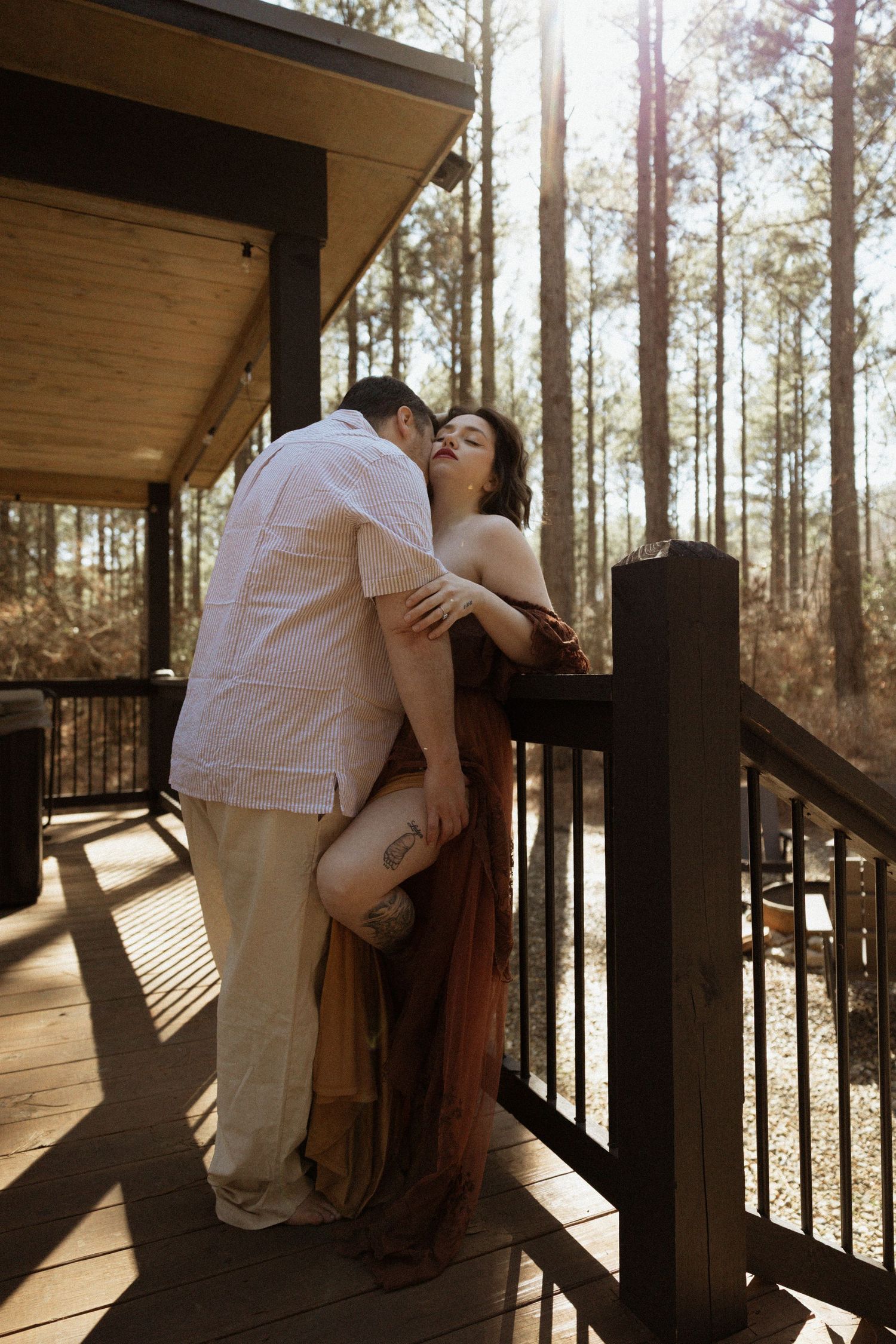 Couple sharing a romantic kiss on a sunlit wooden deck surrounded by tall pine trees.
