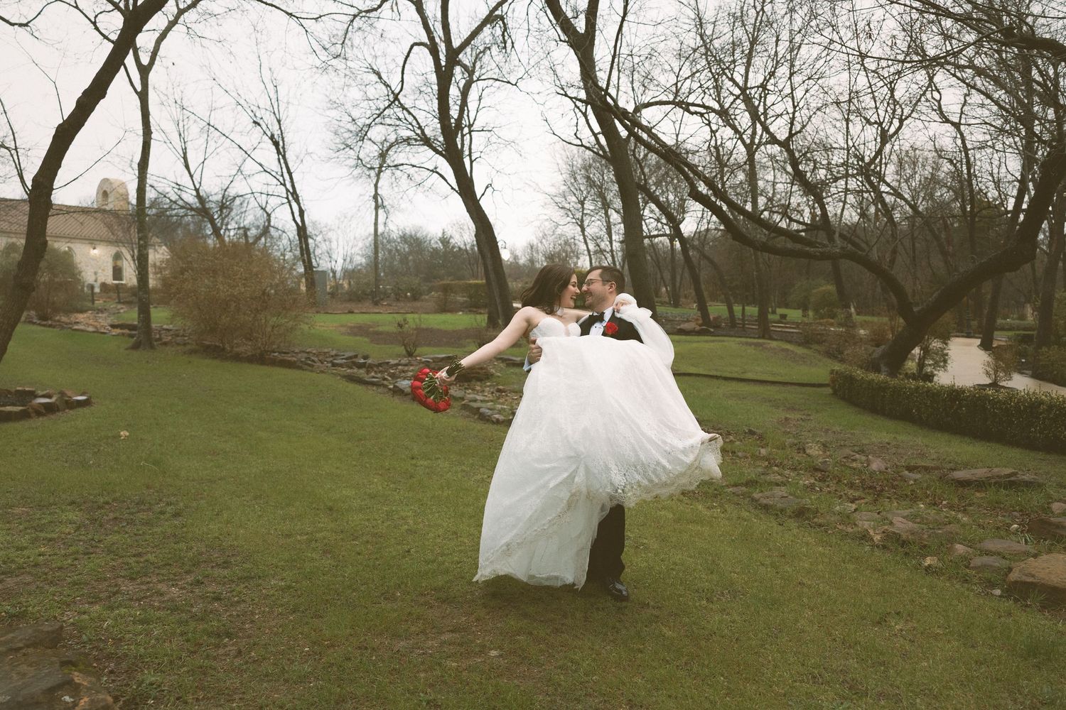 Groom lifting bride in white wedding dress outdoors in a park setting.