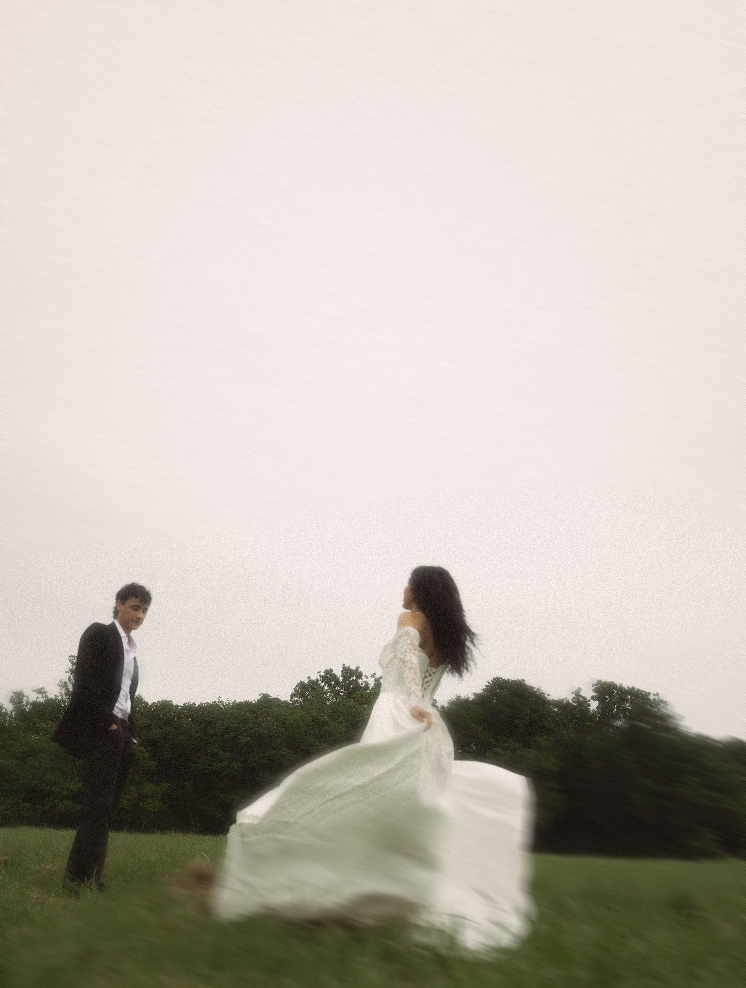 Bride in flowing white gown running toward groom in black suit on a misty green field.
