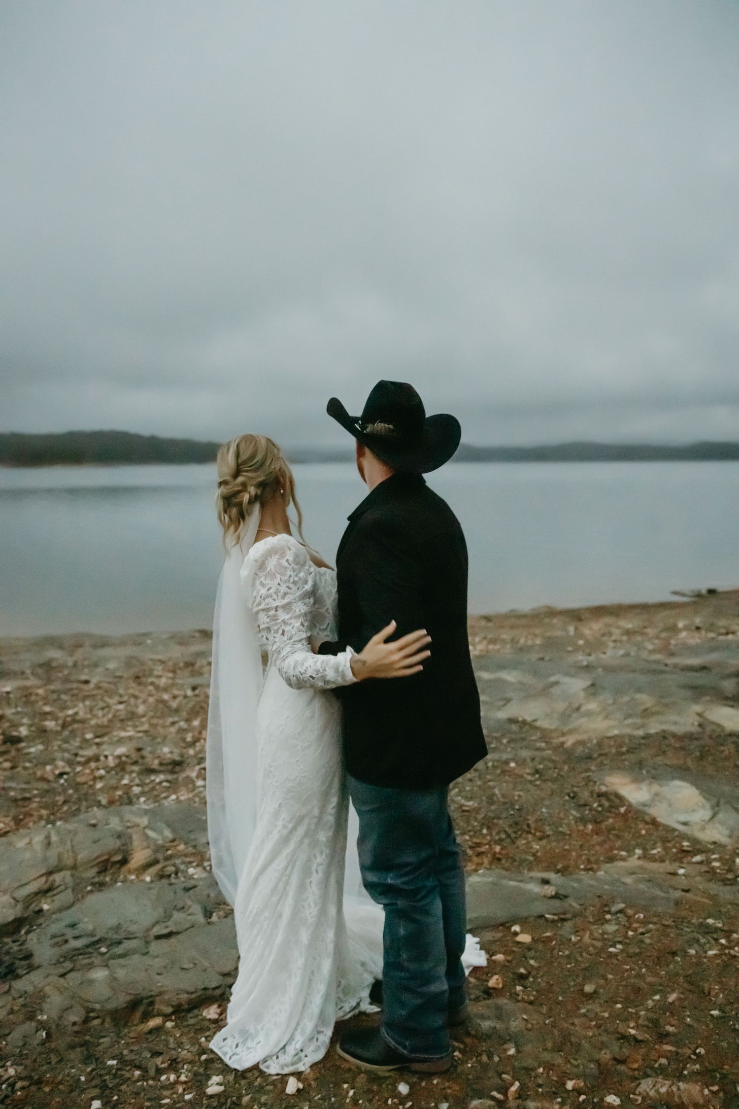 Bride in lace gown and groom in cowboy hat standing together overlooking a calm lake.