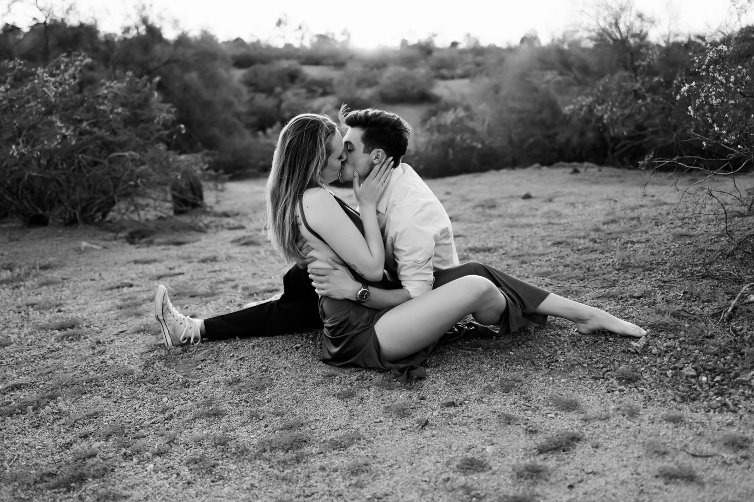 Black and white photo of a couple kissing while sitting on dry desert ground.