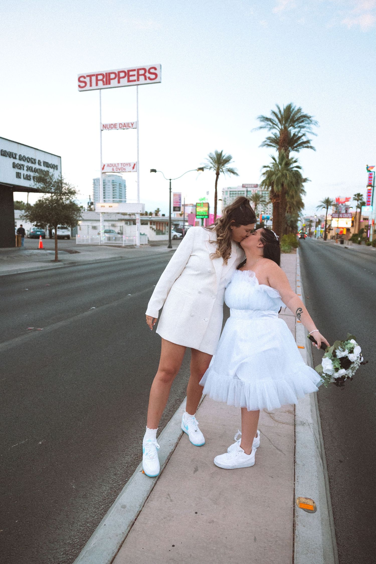 Two brides in white outfits share a kiss on a Las Vegas sidewalk with a Strippers sign behind them.