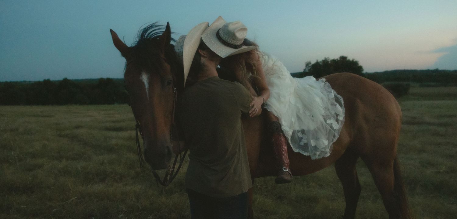 Bride in white dress and groom in cowboy hat stand beside a chestnut horse in an open field at dusk.