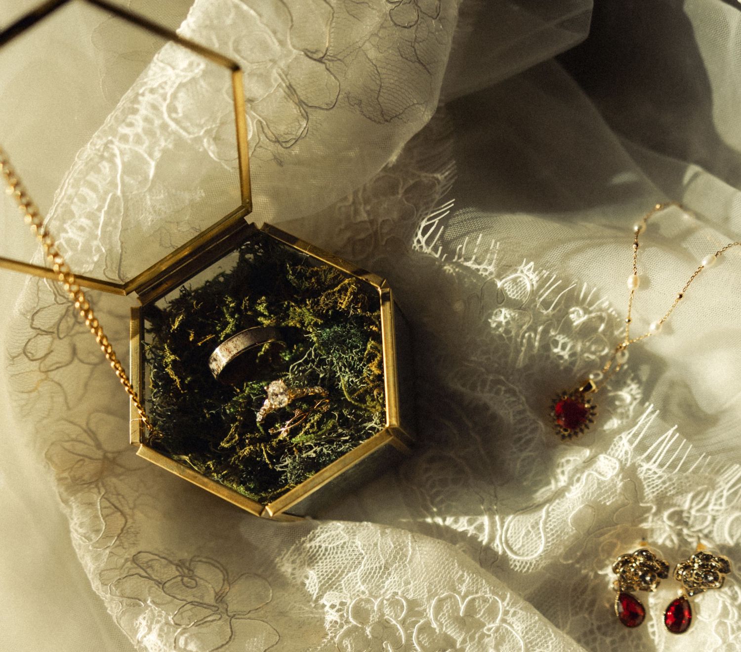 Gold hexagonal glass ring box with moss holds wedding bands beside ruby earrings on white lace fabric.