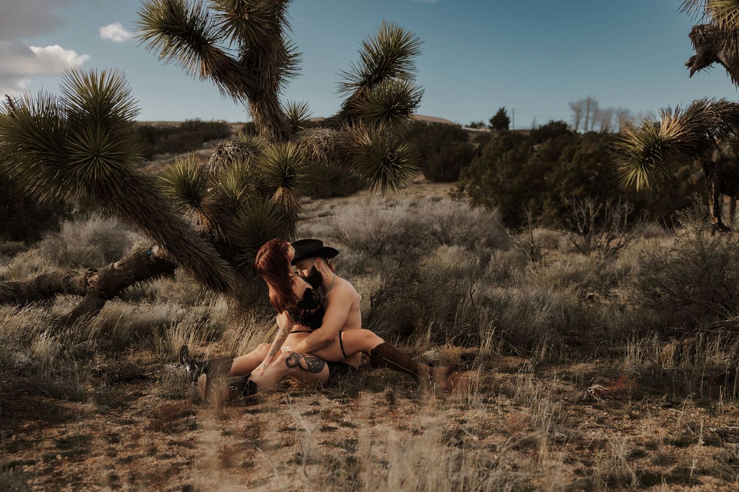 Two people share an intimate embrace seated beneath a Joshua tree in a desert landscape setting.
