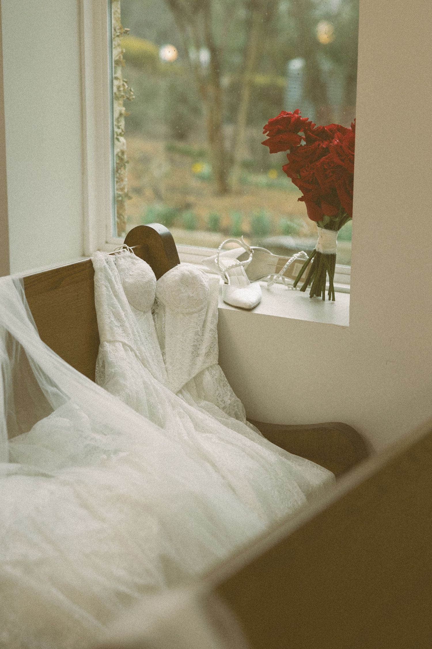 A white lace wedding dress, heels, and red roses arranged by a sunlit window in a warm-toned room.