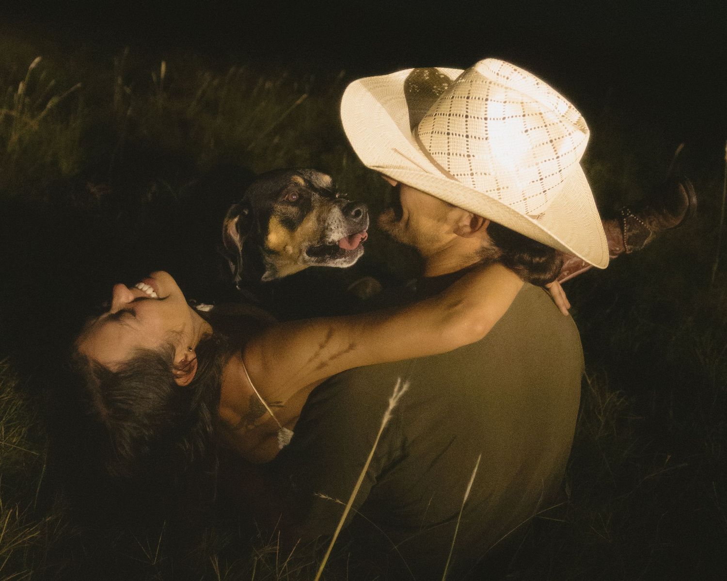 Couple cuddling with small dog in dark grassy field at night.