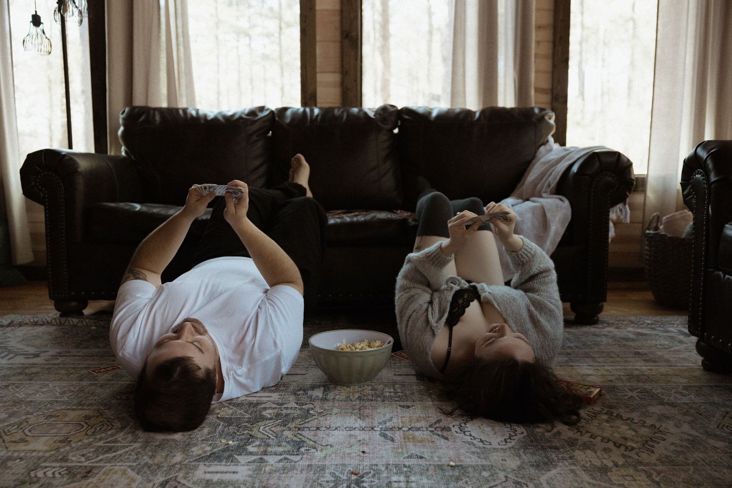 Couple lying on floor using smartphones with snacks between them near dark leather sofa.
