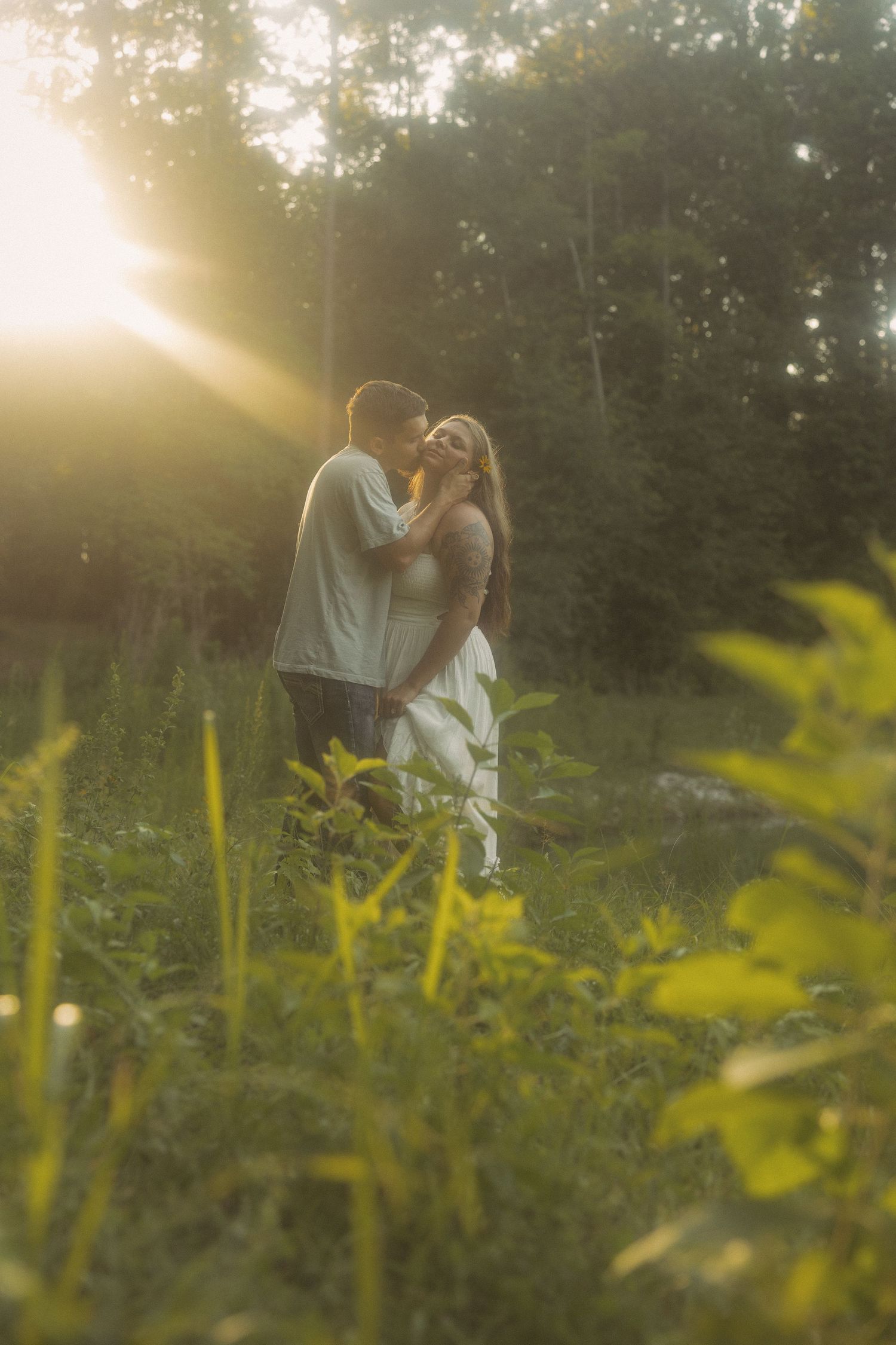 A couple embraces in a sunlit meadow surrounded by green foliage during golden hour.