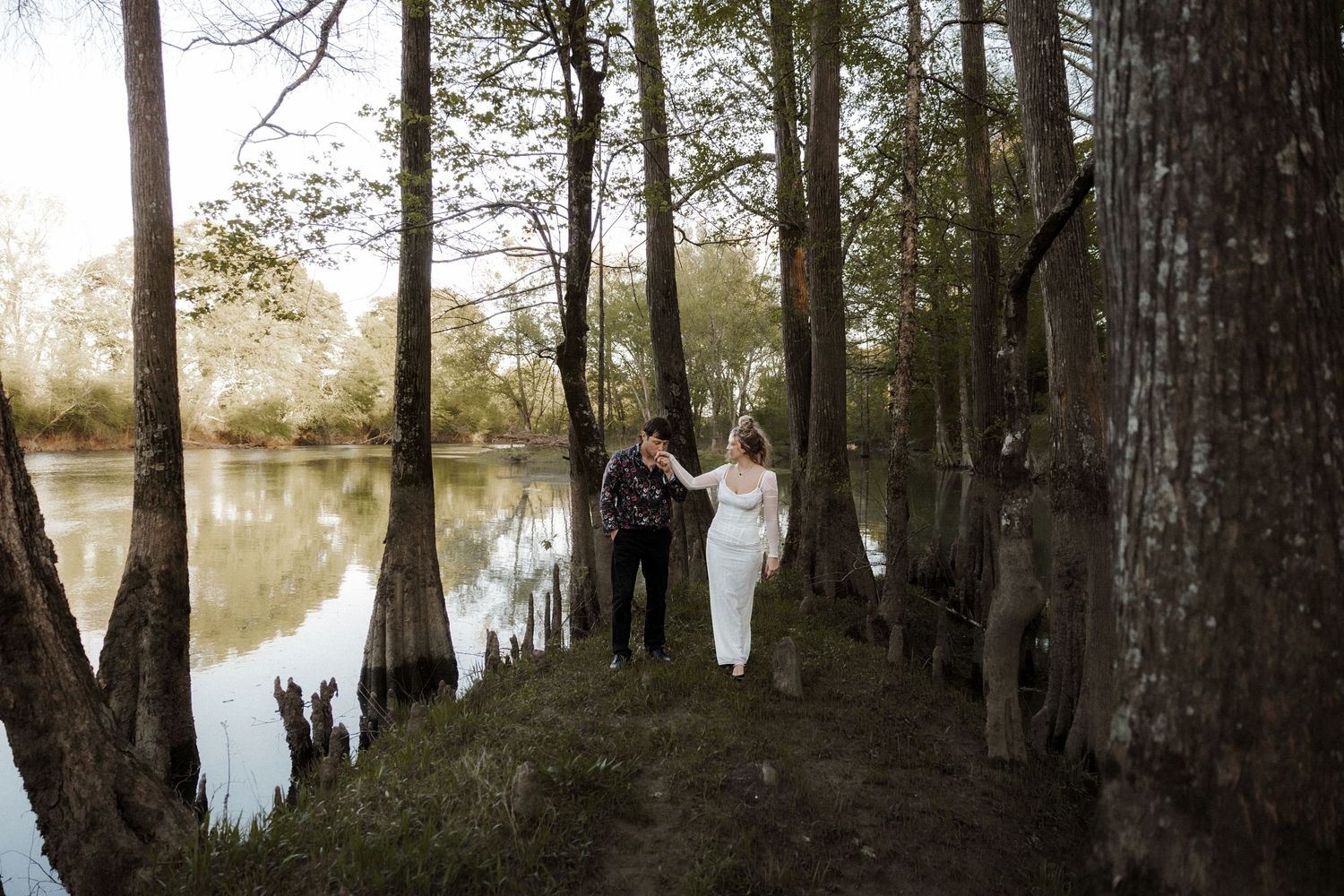 A couple walks hand in hand among cypress trees beside a sunlit river in a romantic outdoor setting.