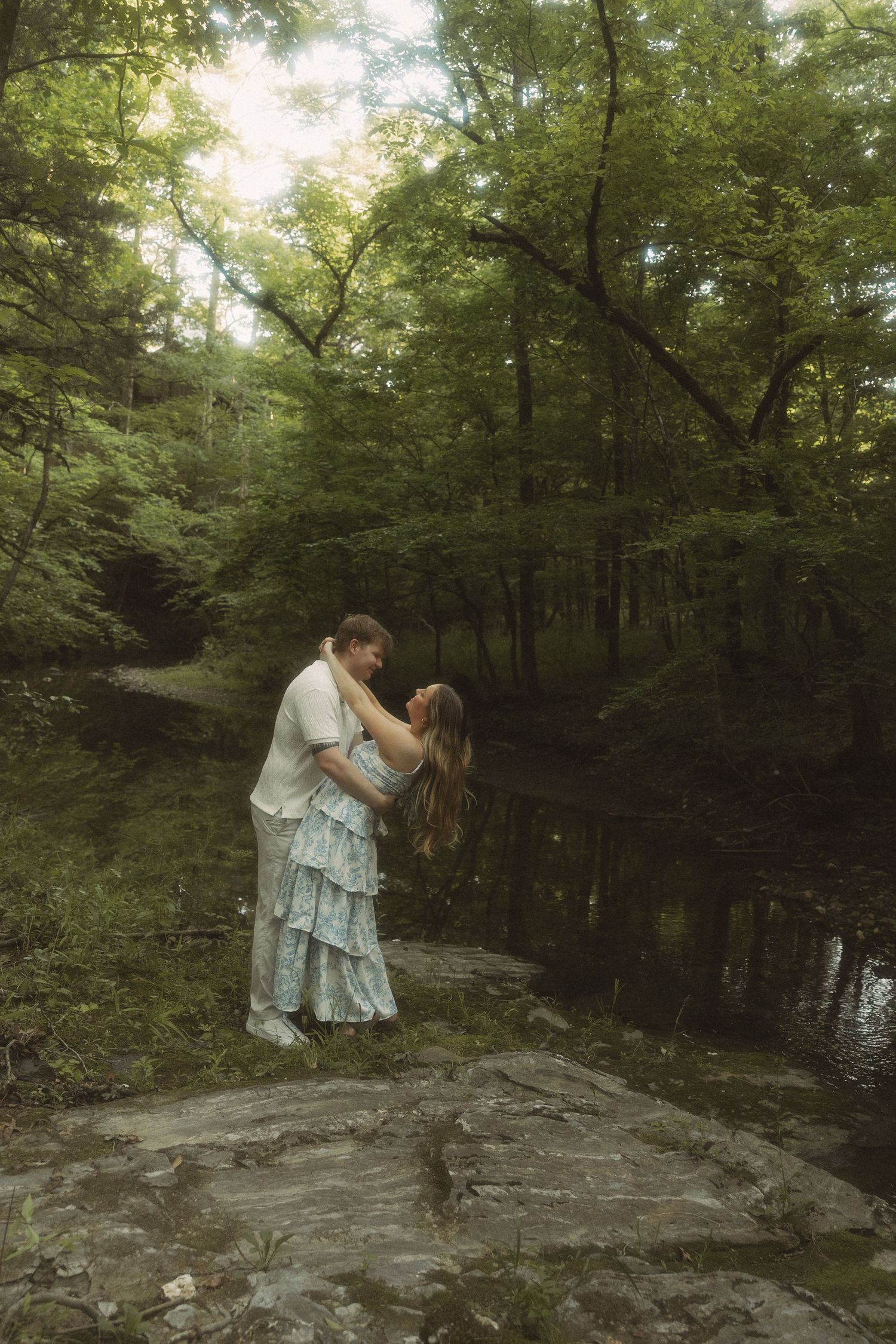 Romantic couple embracing near a serene forest creek surrounded by lush green trees.