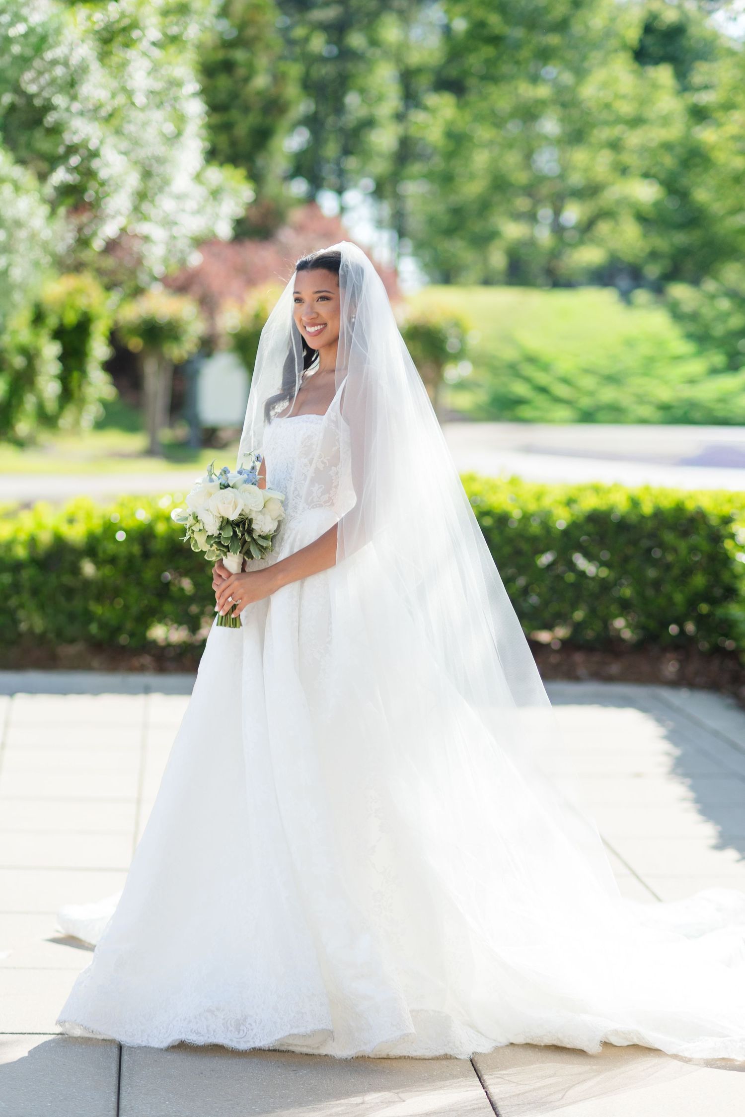 Smiling bride in white lace gown and veil holding bouquet of white roses outdoors on sunny day.