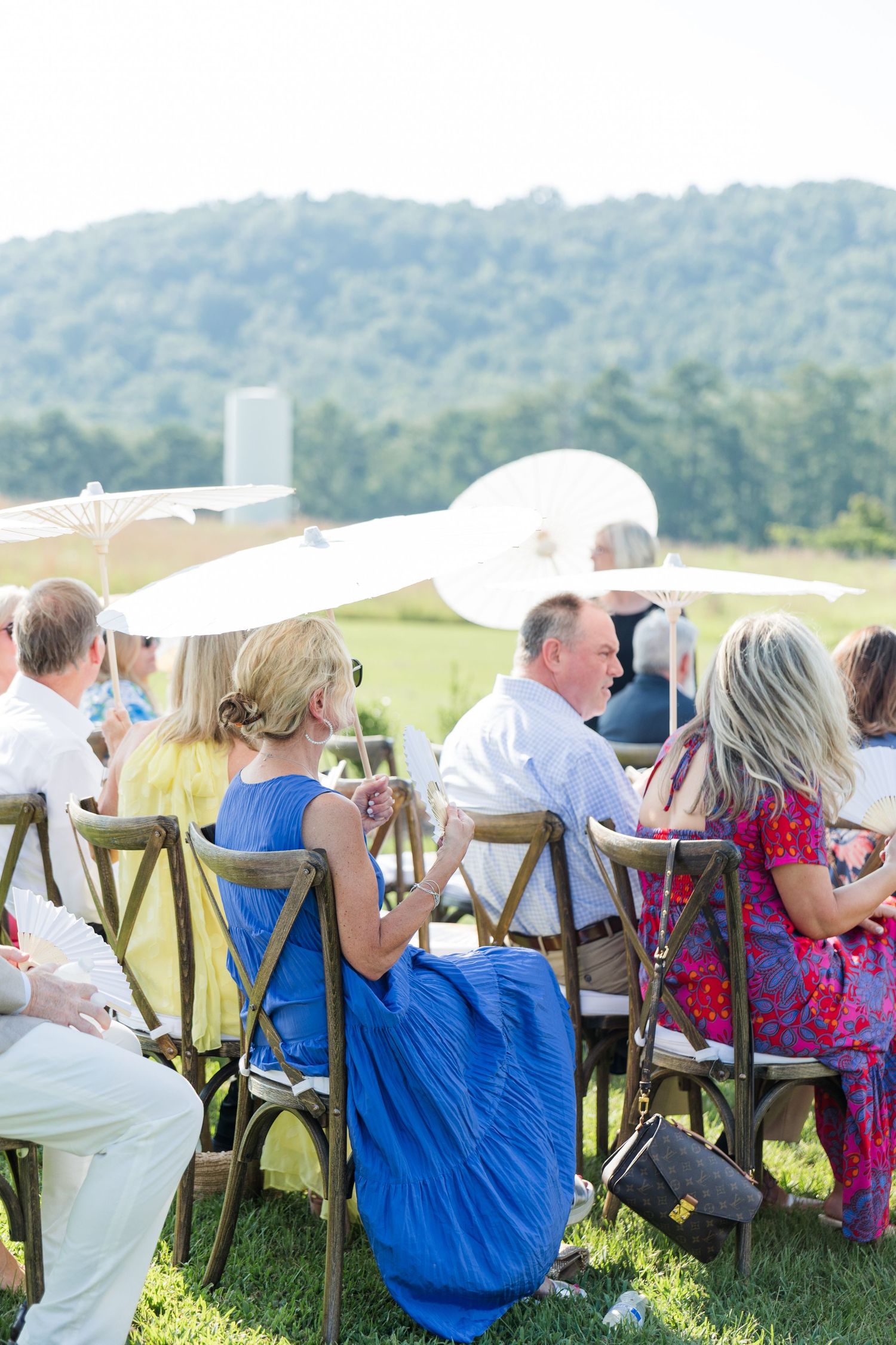 Guests seated outdoors in wooden chairs under white parasols at a sunny garden event with rolling hills in the background.