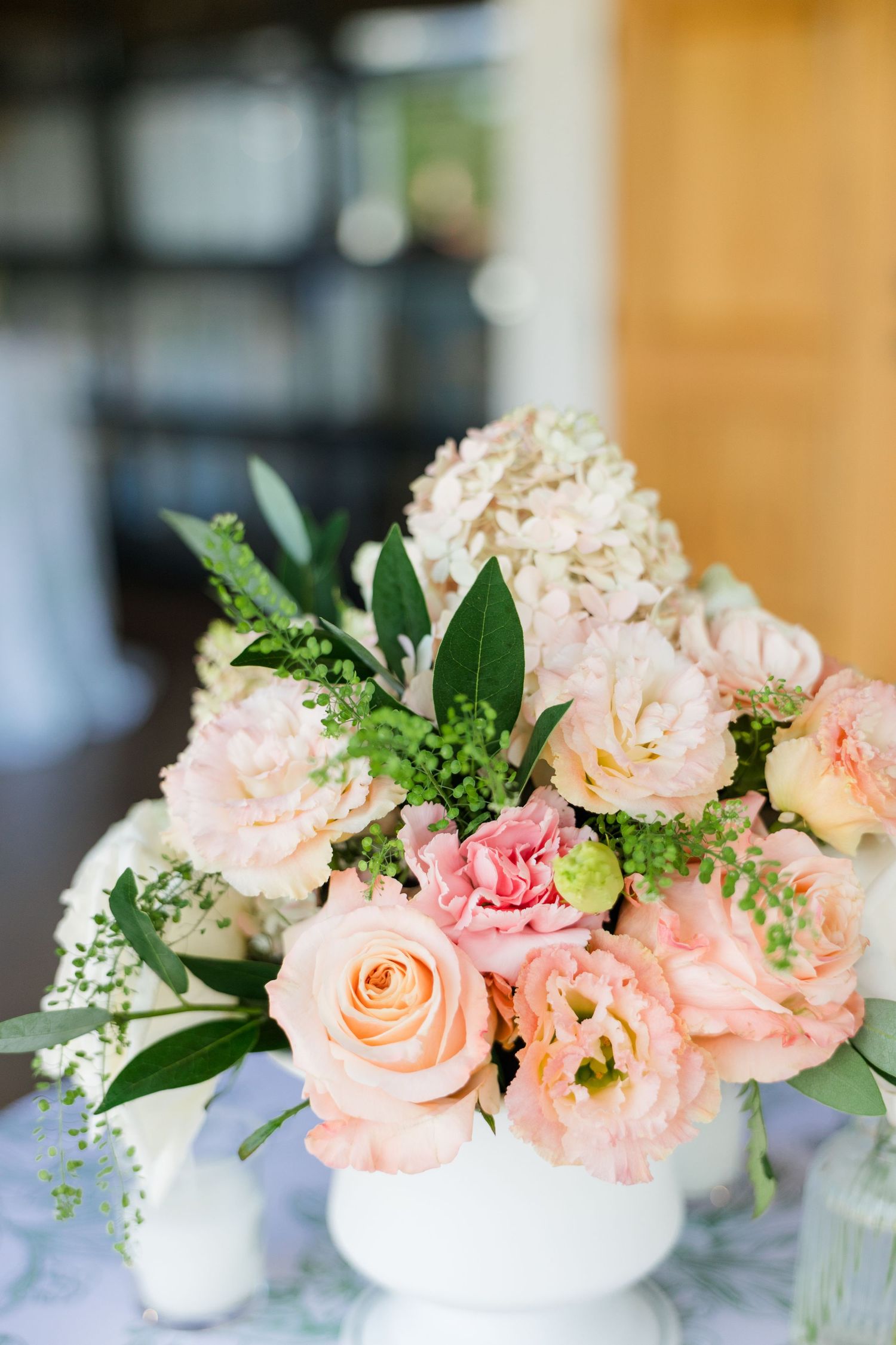 White vase with blush pink roses, peonies, and hydrangeas centerpiece arrangement with green foliage.