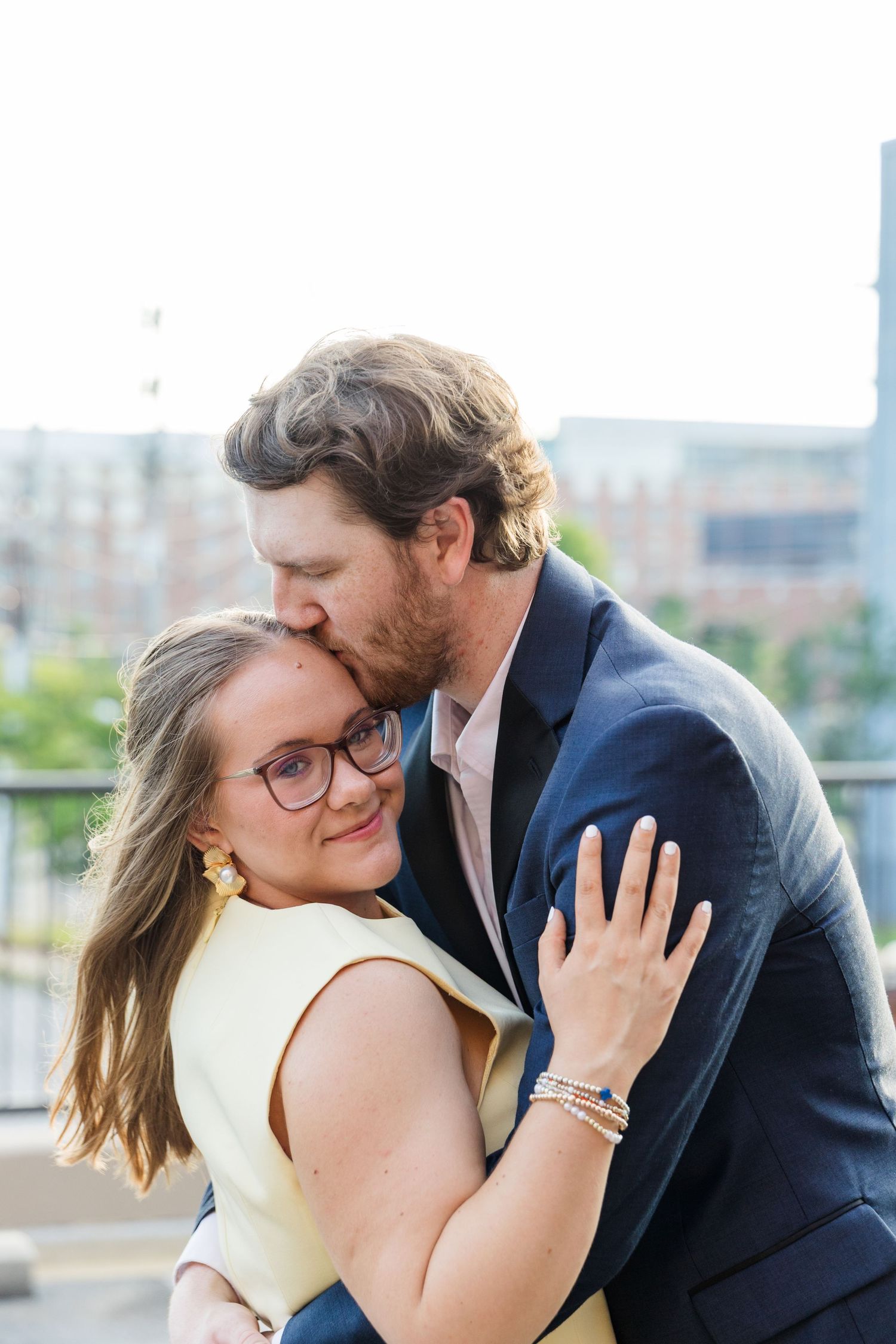 A couple shares a tender moment, man in navy suit kissing woman in yellow dress on the forehead outdoors.