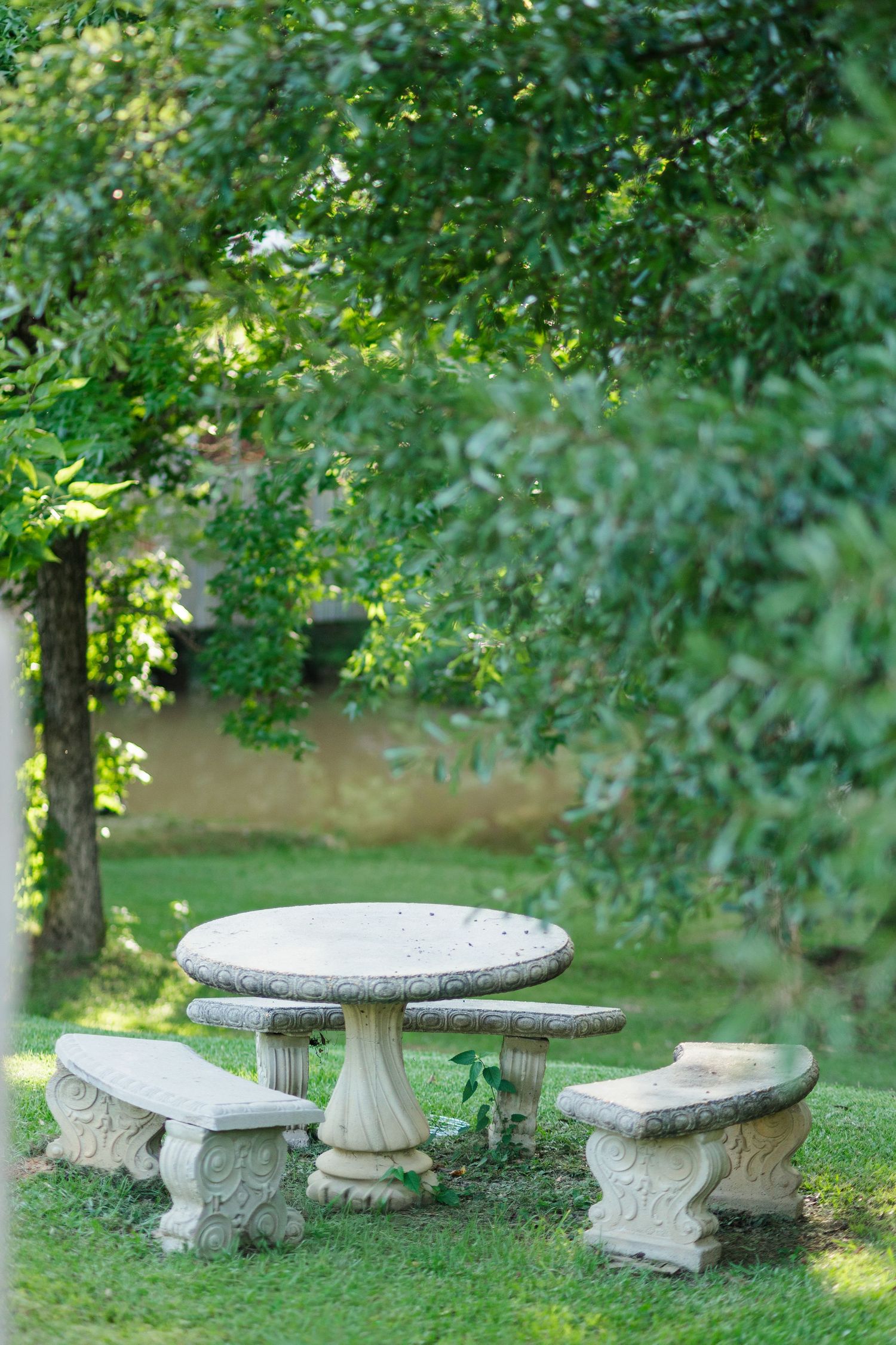Stone garden table and benches set on green lawn surrounded by lush trees in a peaceful outdoor setting.
