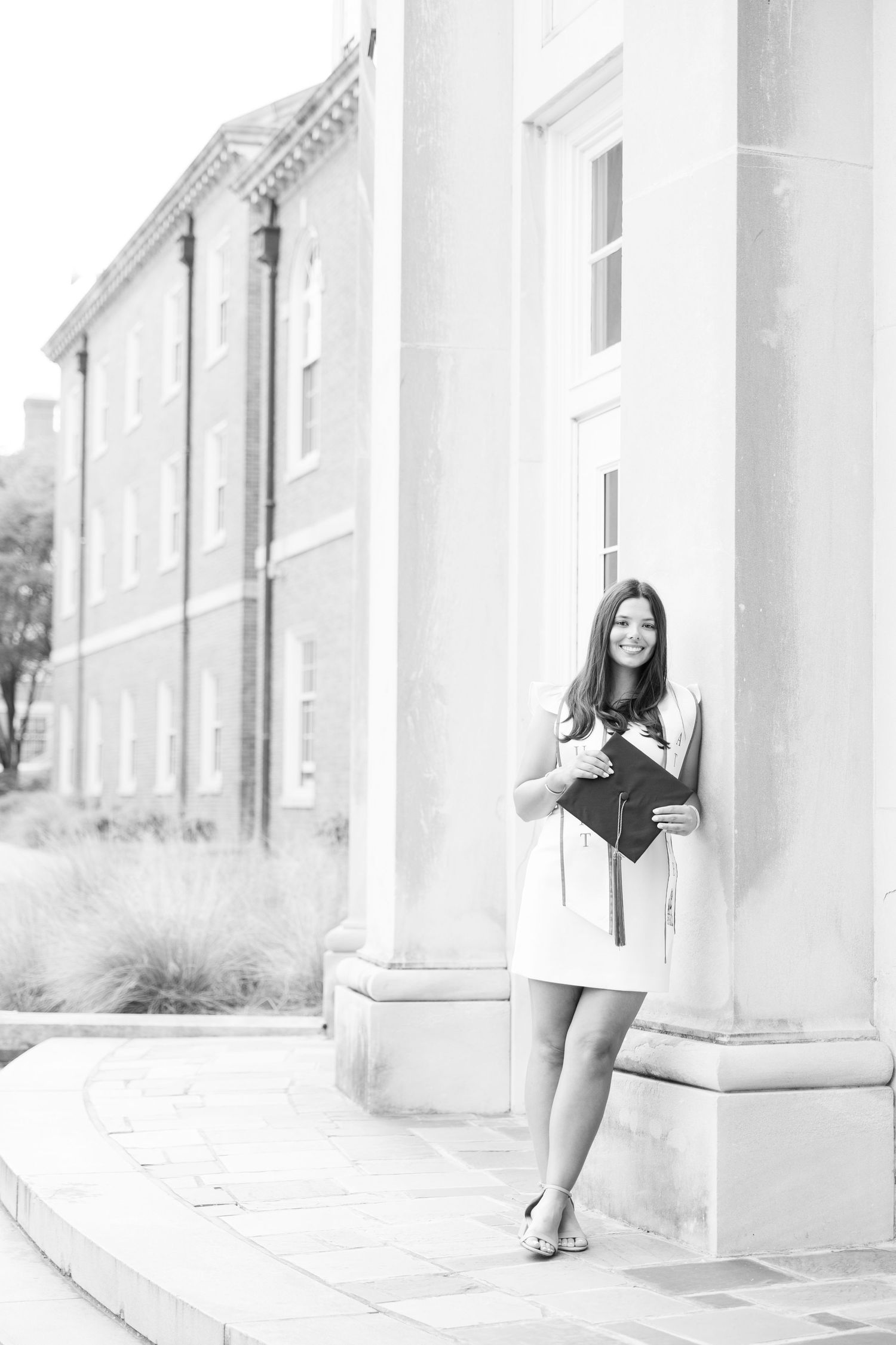 Young woman in white dress holding graduation cap leans against column of brick university building in black and white photo.