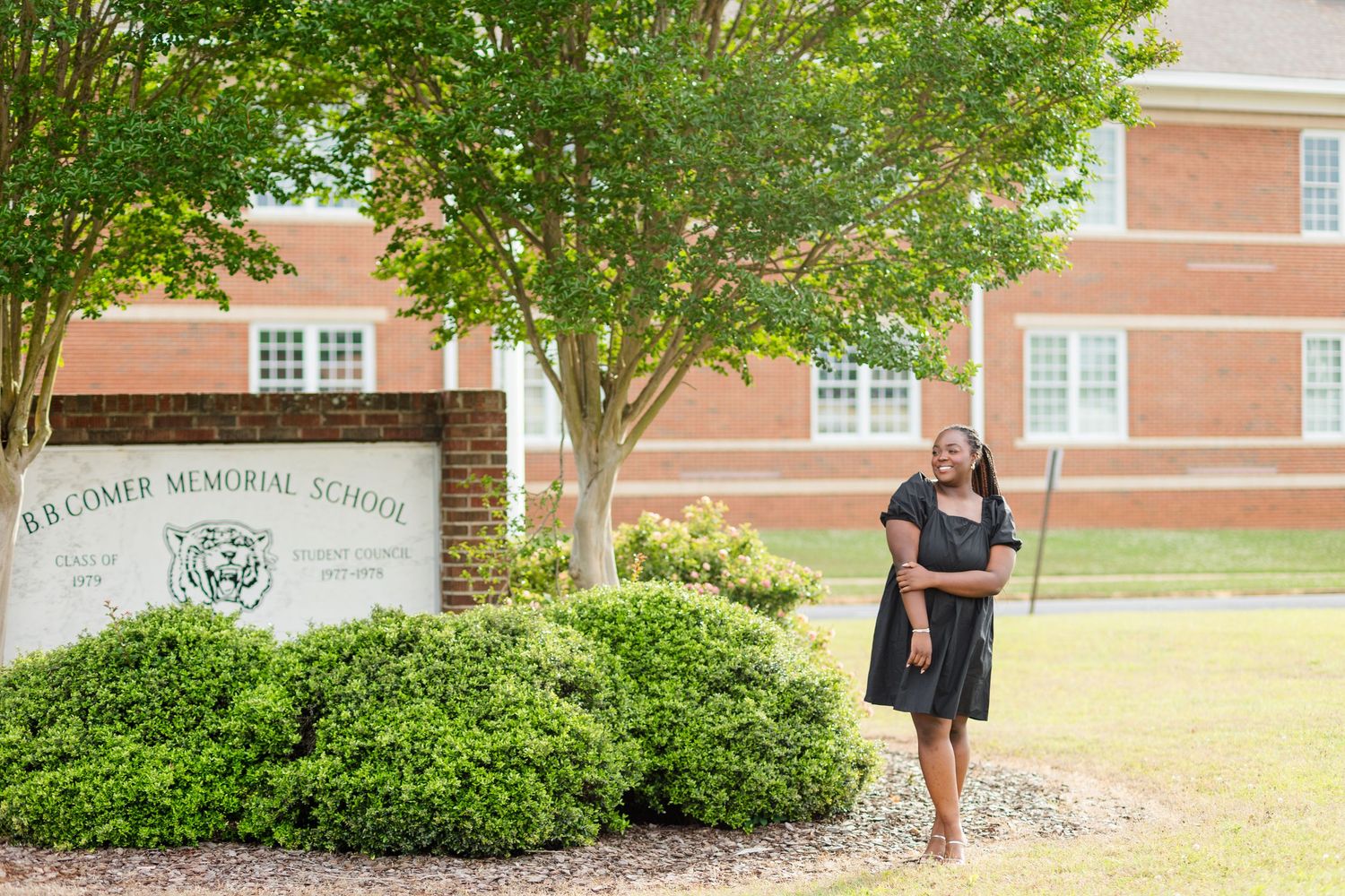 A young woman in a black dress poses near B.B. Comer Memorial School sign surrounded by lush green trees.