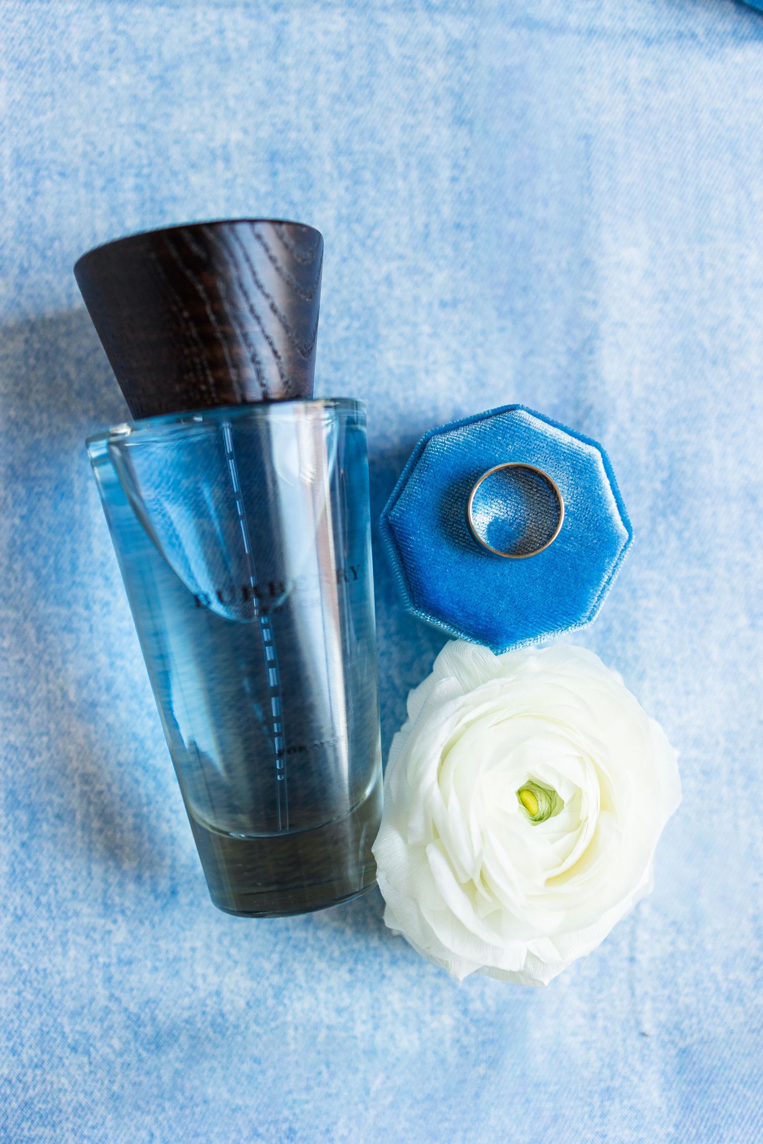 Blue perfume bottle with wooden cap beside a blue velvet ring box with gold ring and white ranunculus flower.