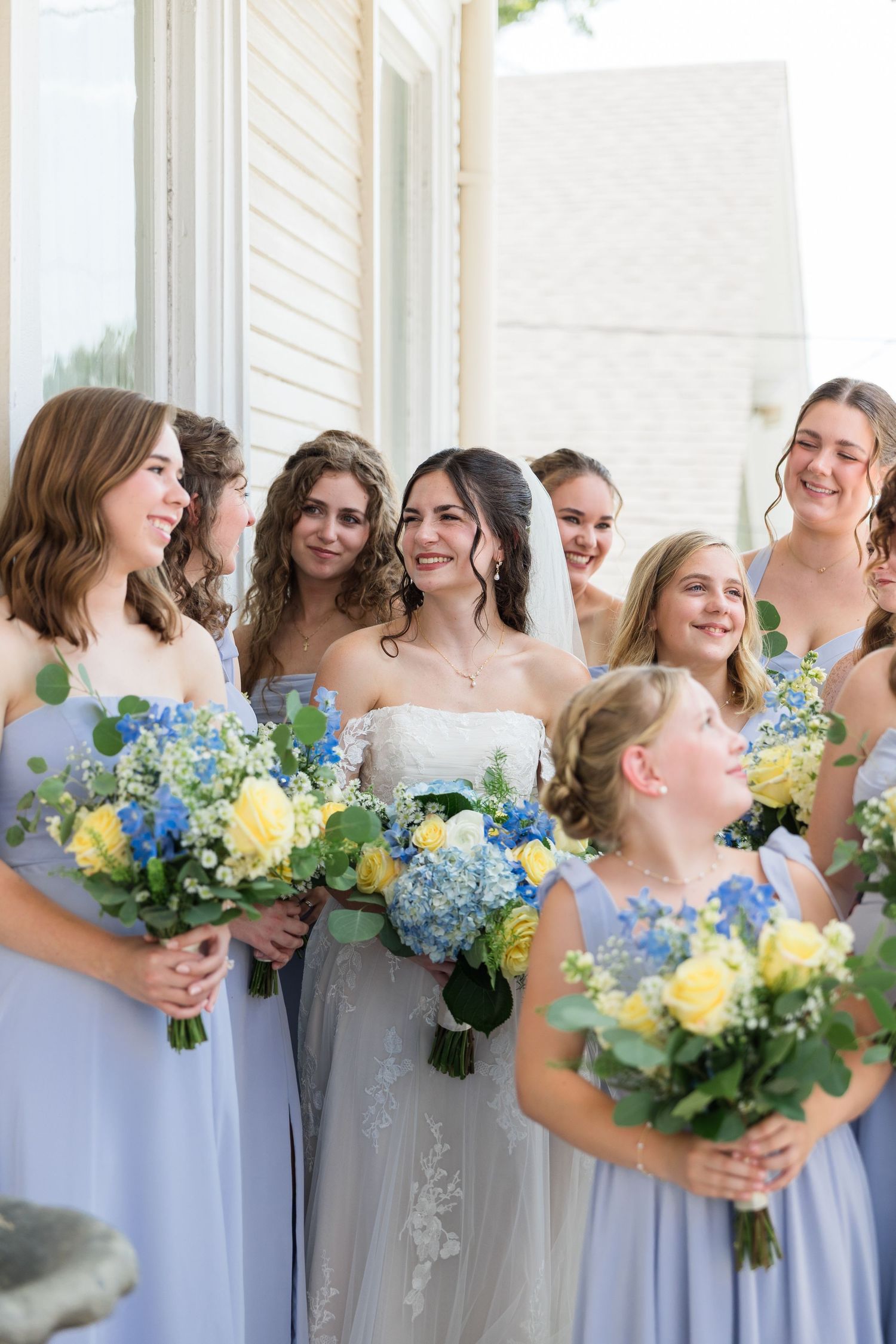 Bride with bridesmaids in lavender dresses holding blue and yellow floral bouquets.