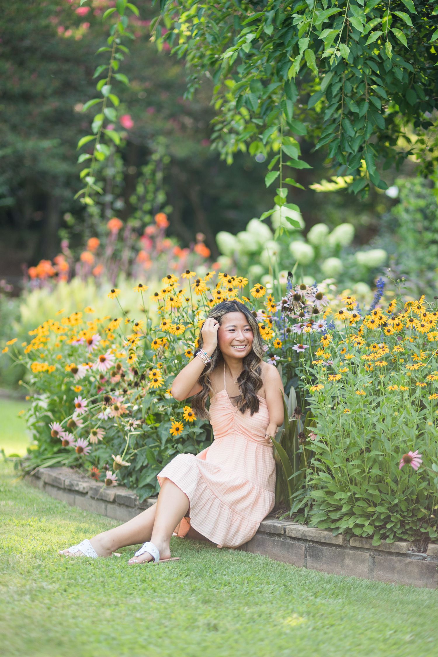 Young woman in peach dress sitting by colorful garden flowers, smiling outdoors.